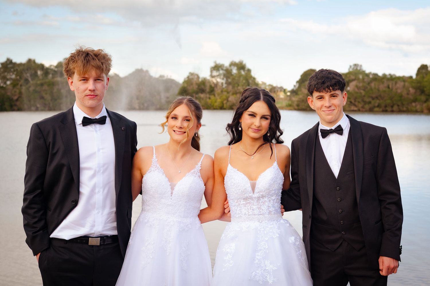 Two couples in formal wear, girls in white gowns and boys in black tuxedos, posing by a lake.