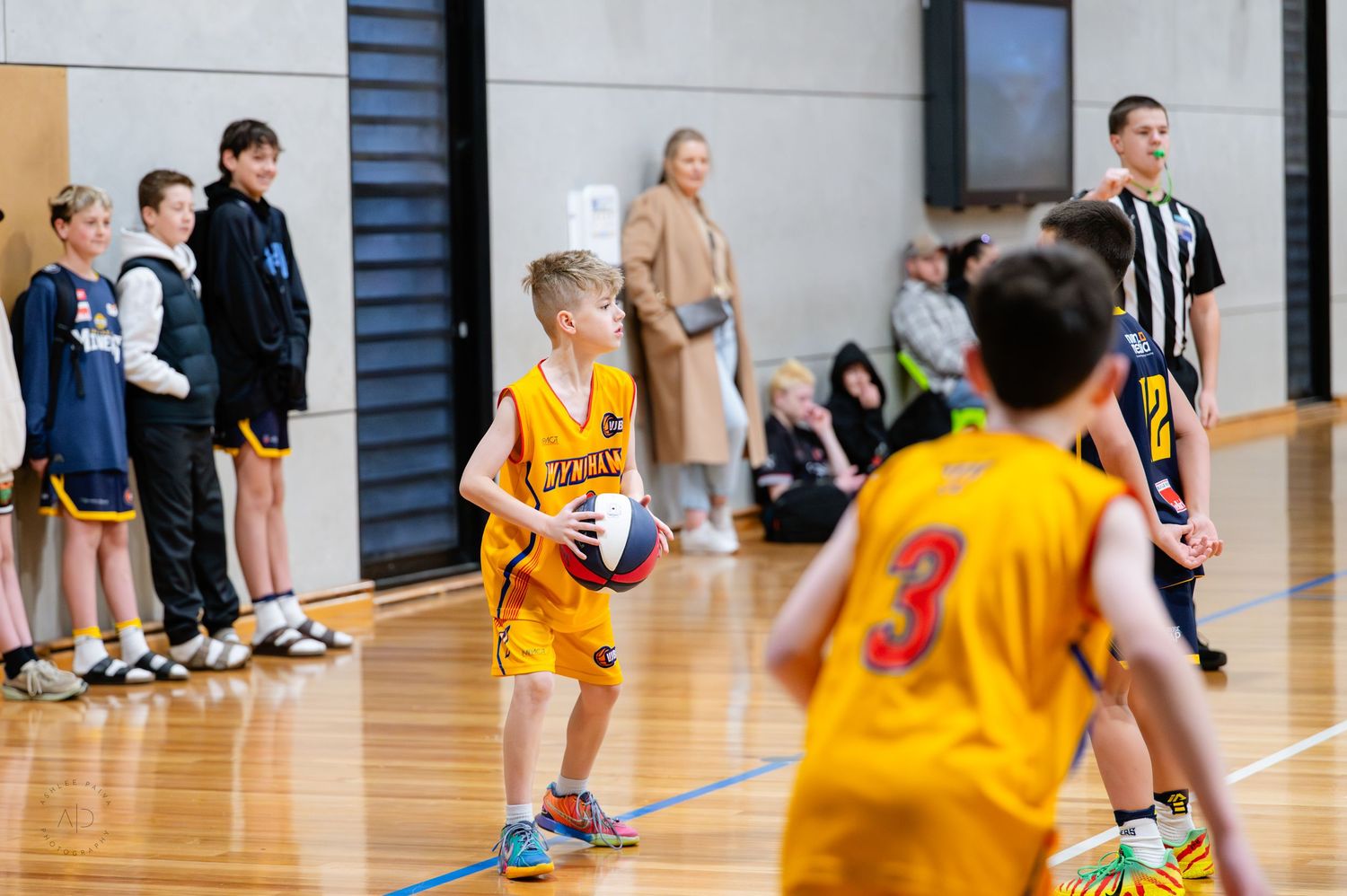 Young boys in yellow uniforms playing an indoor basketball game on a polished court with spectators watching.