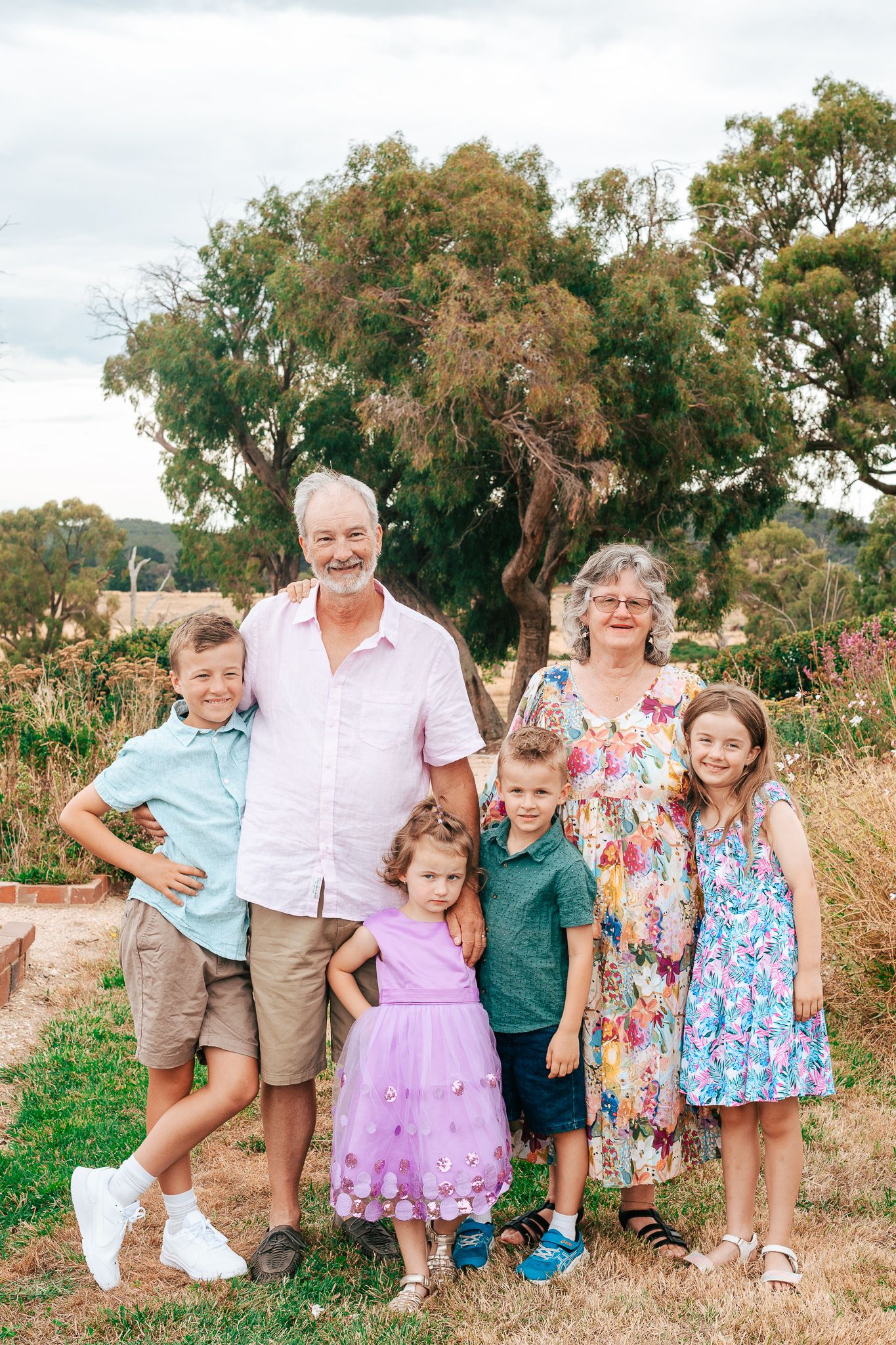 A multi-generational family poses together outdoors in casual summer attire on a grassy hillside.