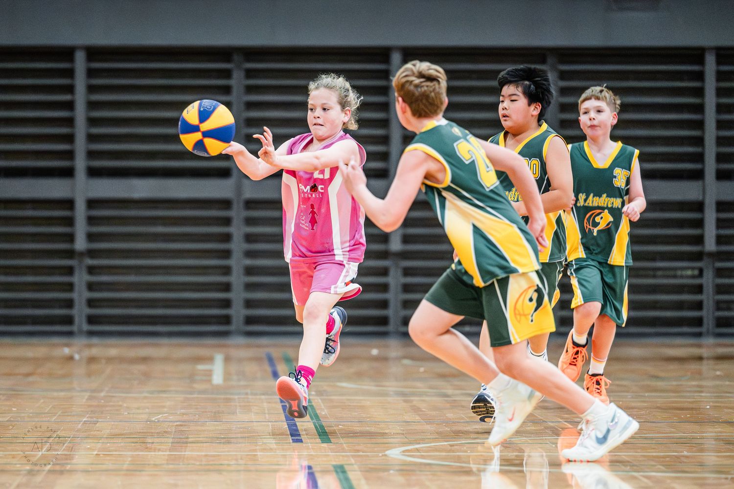 Young girl in pink uniform passing basketball during youth indoor basketball game against green and gold team.