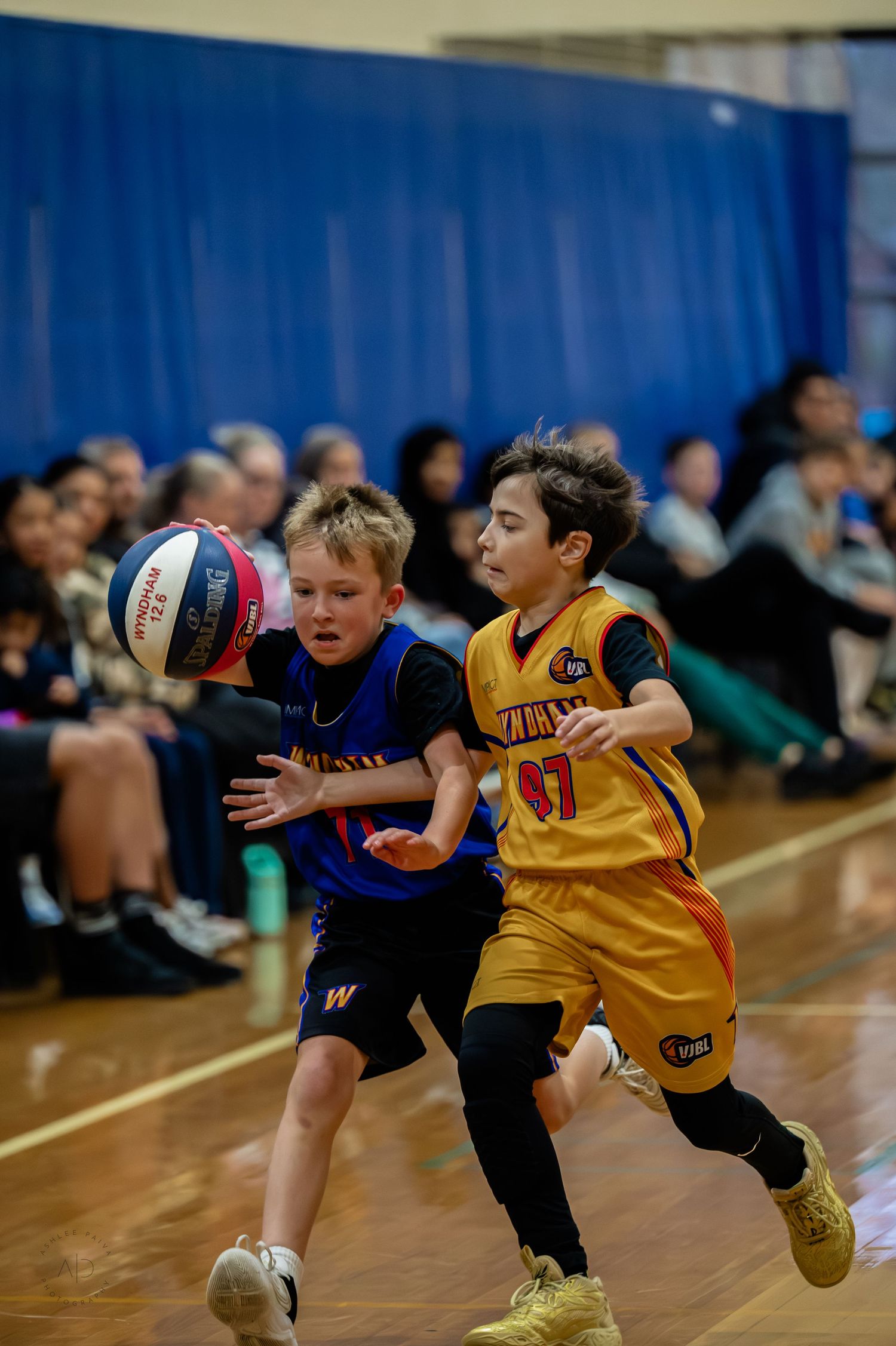 Two young boys compete in a youth basketball game, one dribbling past a defender in a packed indoor gymnasium.