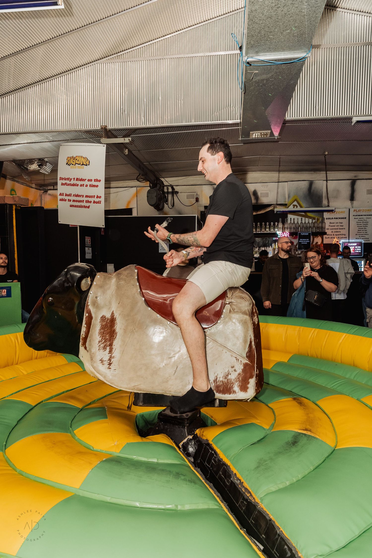 Man riding a mechanical bull at an indoor fairground attraction, surrounded by spectators watching the rodeo challenge.