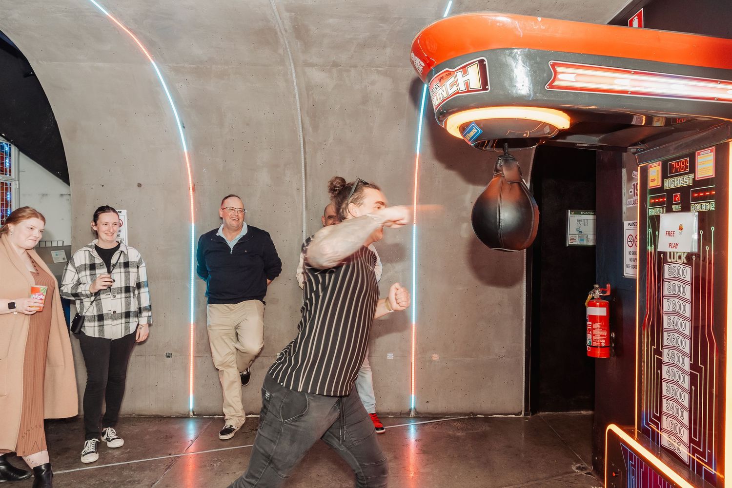 Person punching a speed bag arcade game while onlookers watch in a dimly lit entertainment venue.
