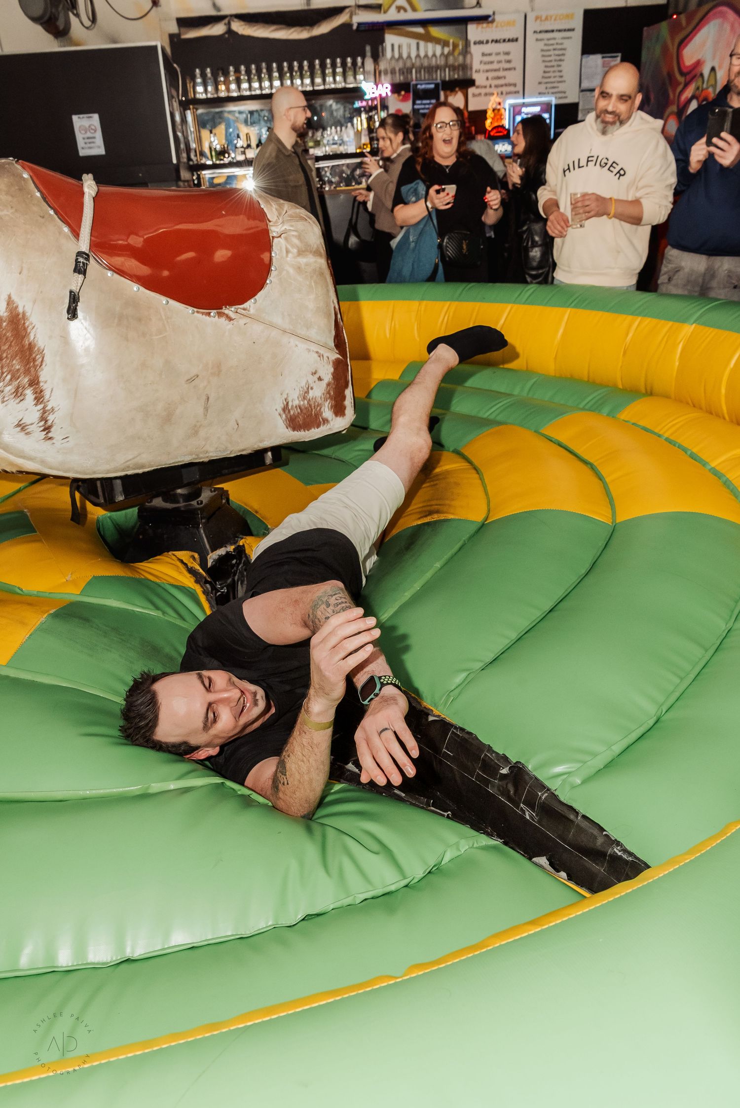 Man fallen off mechanical bull on inflatable mat at indoor bar event with spectators watching and laughing.