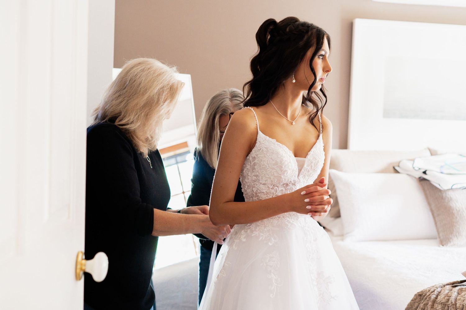 Bride in lace wedding gown being helped by woman in black dress in bright bedroom before ceremony.