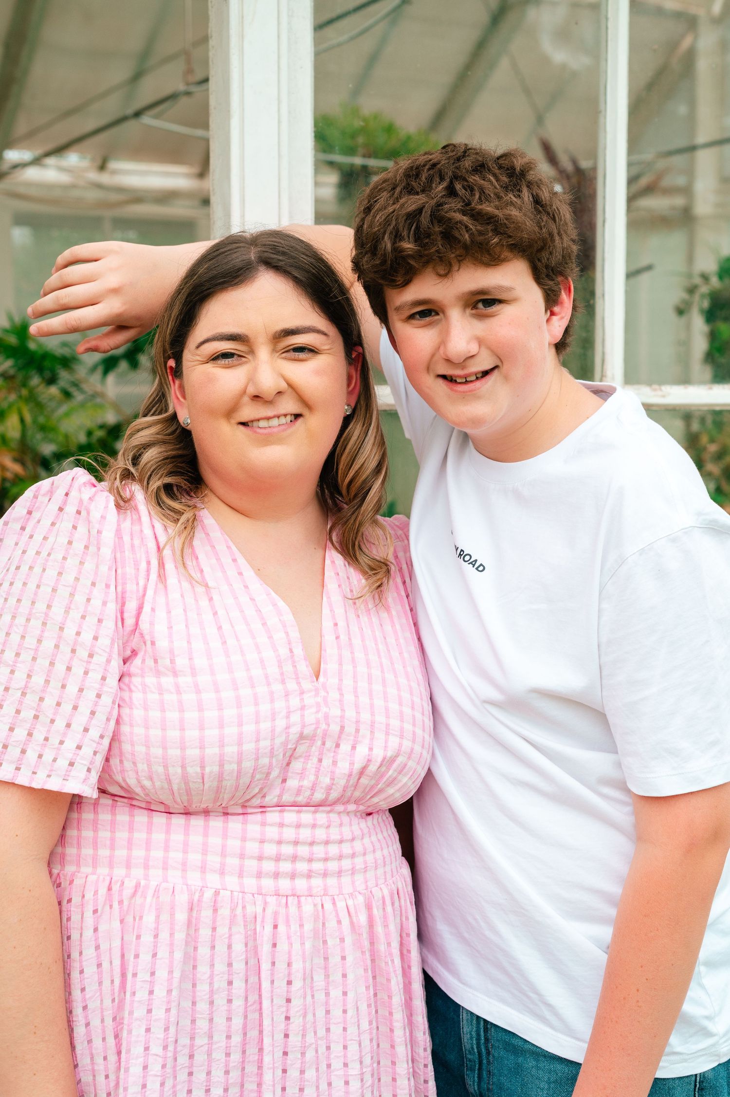 Two people pose together outdoors, one wearing a pink gingham dress and the other in a white t-shirt.