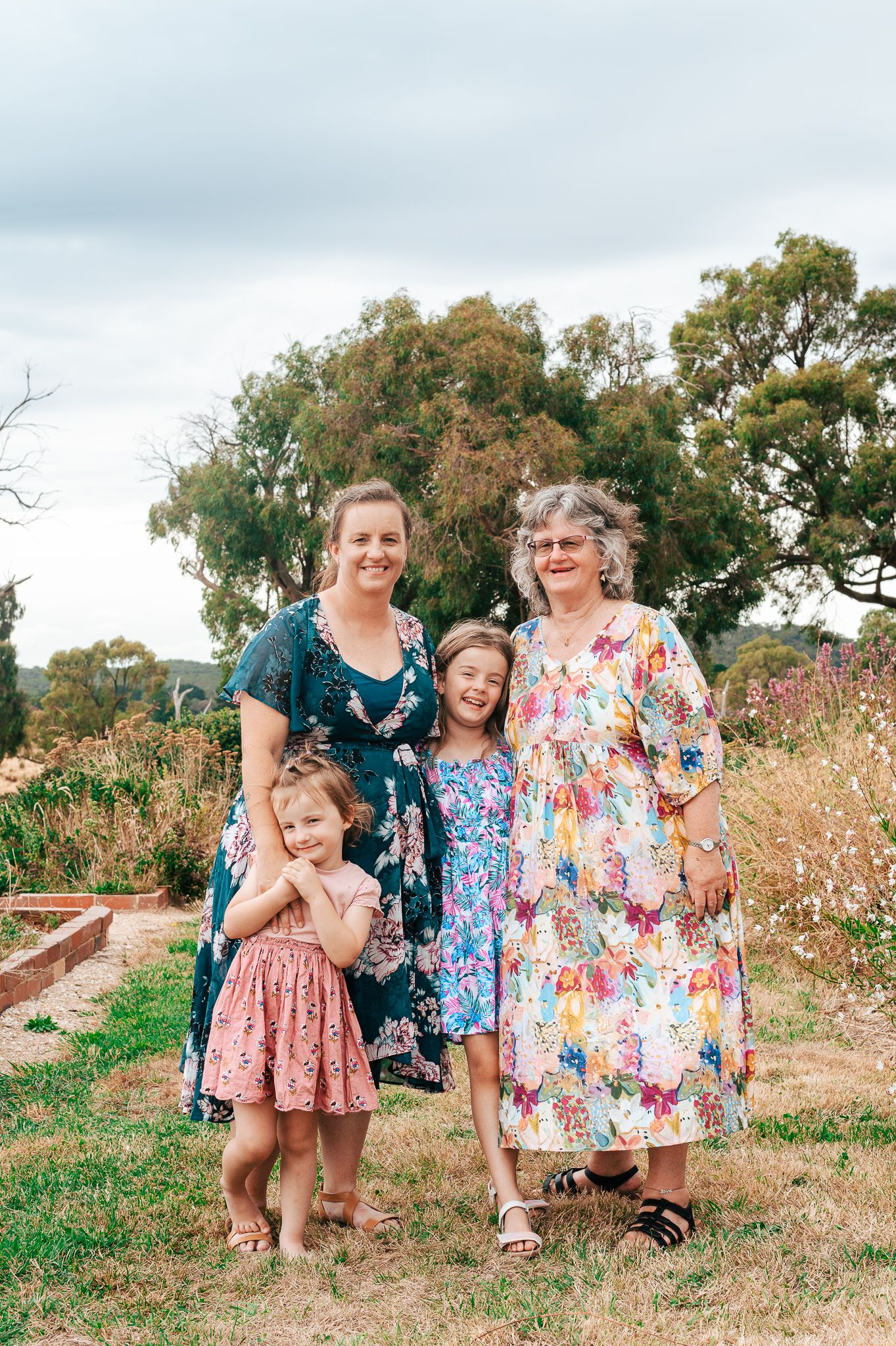 Three generations pose together outdoors in floral dresses on a sunny day with trees and garden beds in the background.
