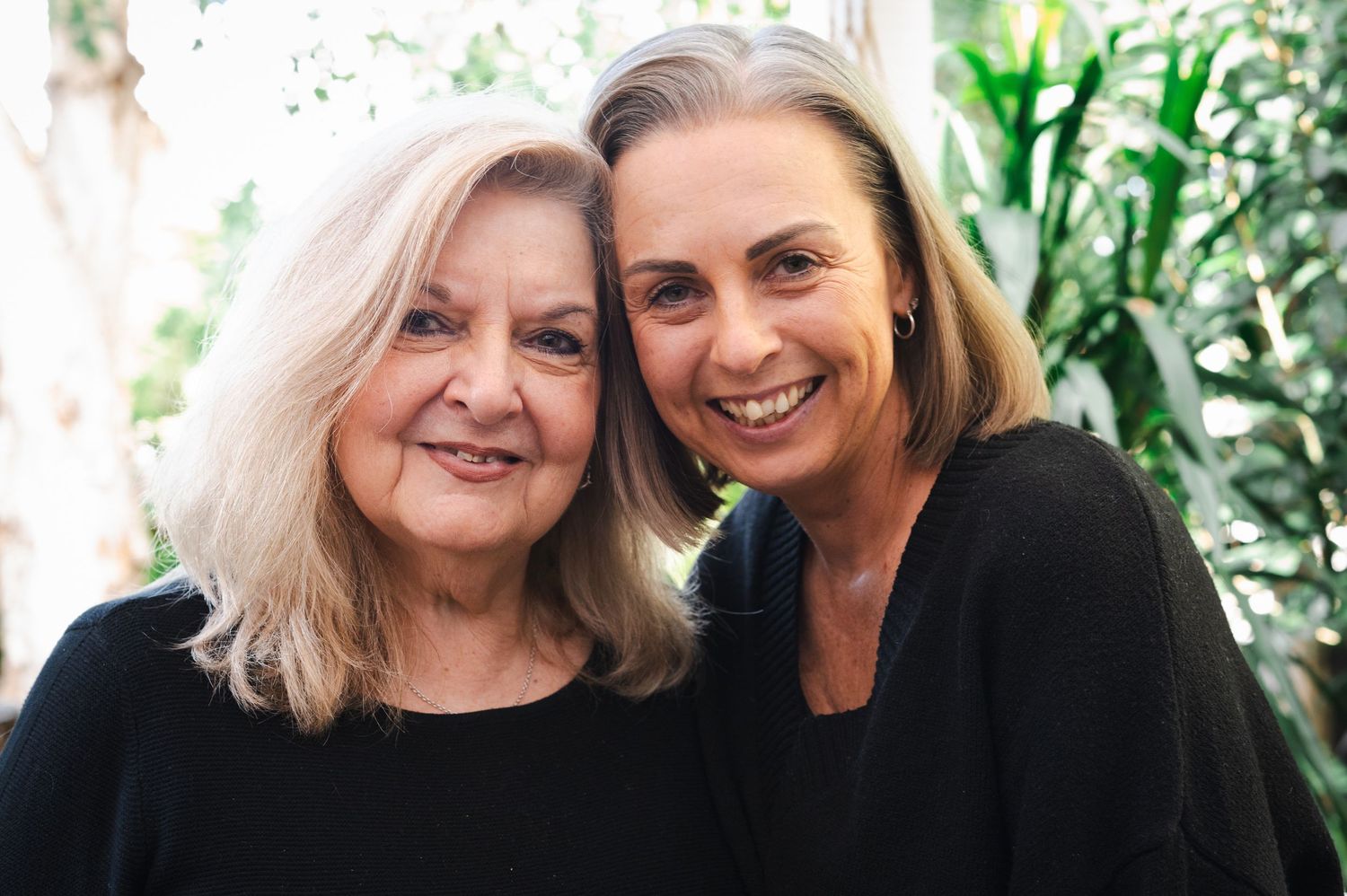 Two smiling women in black tops pose together outdoors near lush green tropical plants.