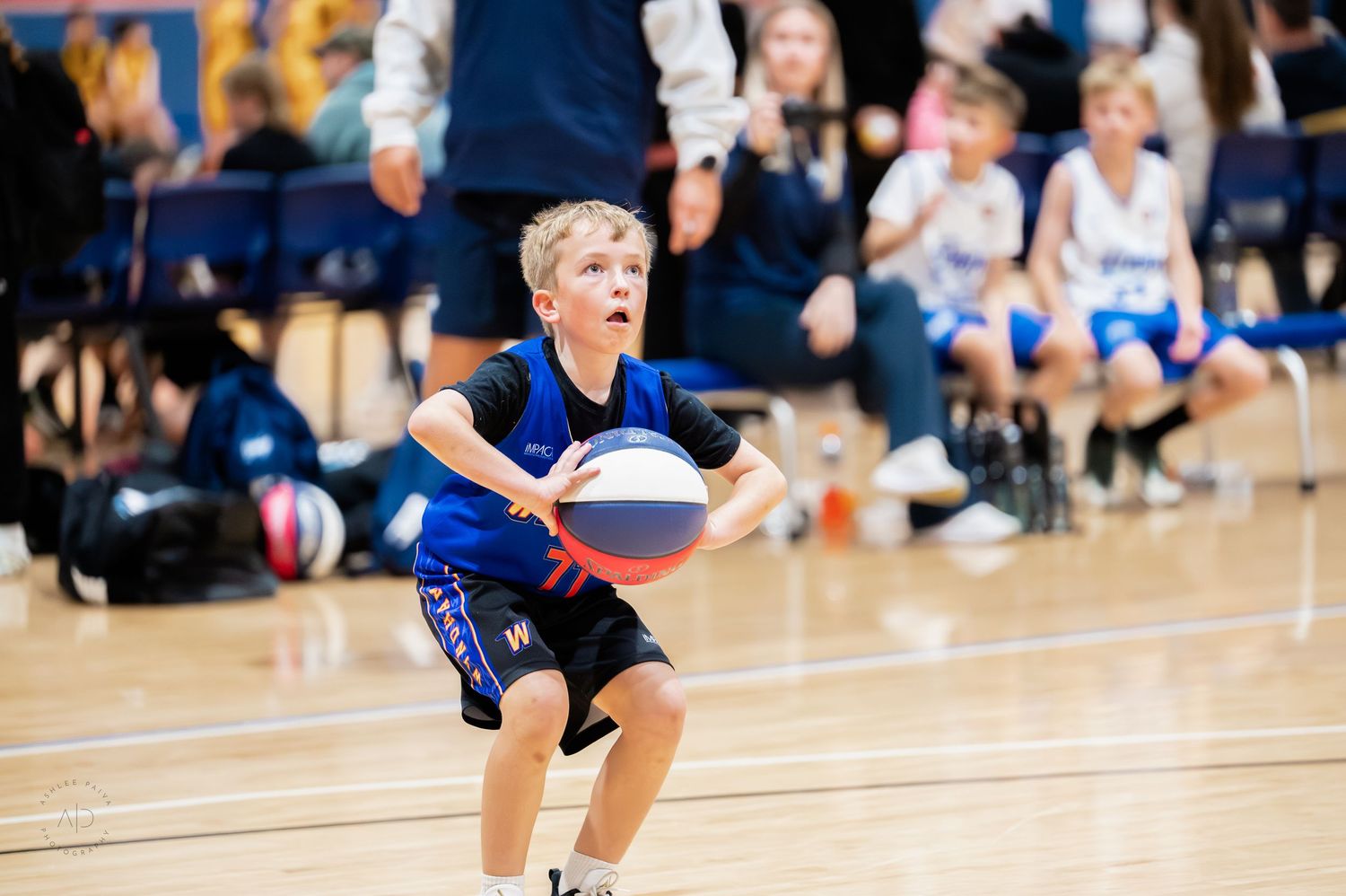 Young boy in blue jersey dribbling a basketball during a youth basketball game on an indoor court.