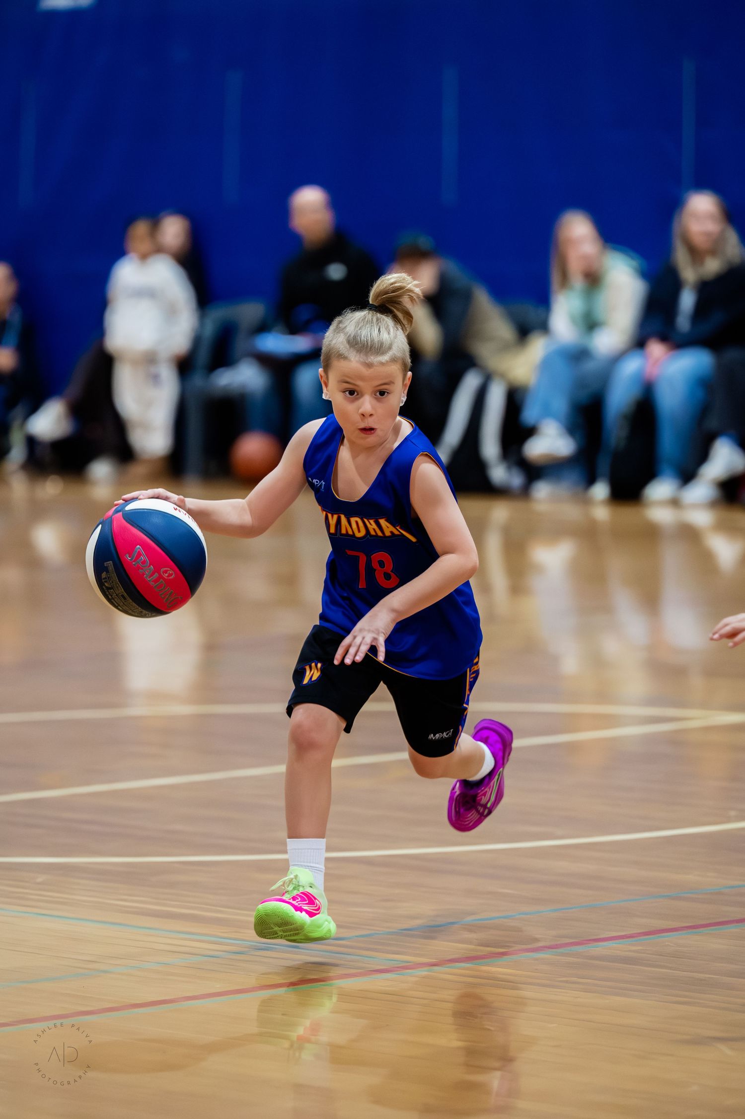 Young girl in blue basketball uniform dribbling ball down court during indoor youth basketball game.