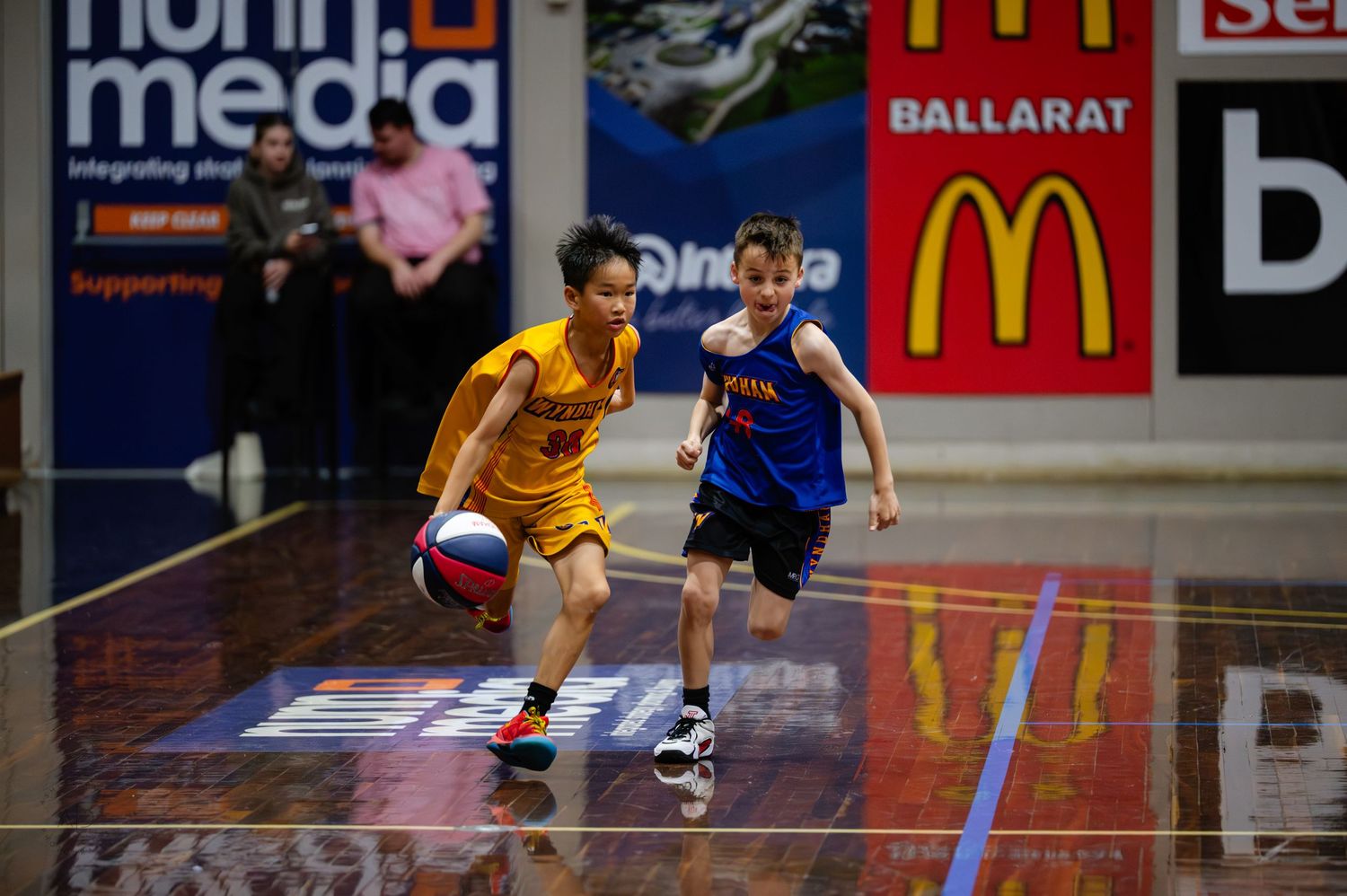 Two young boys playing basketball indoors in Ballarat, one dribbling the ball in a yellow jersey.