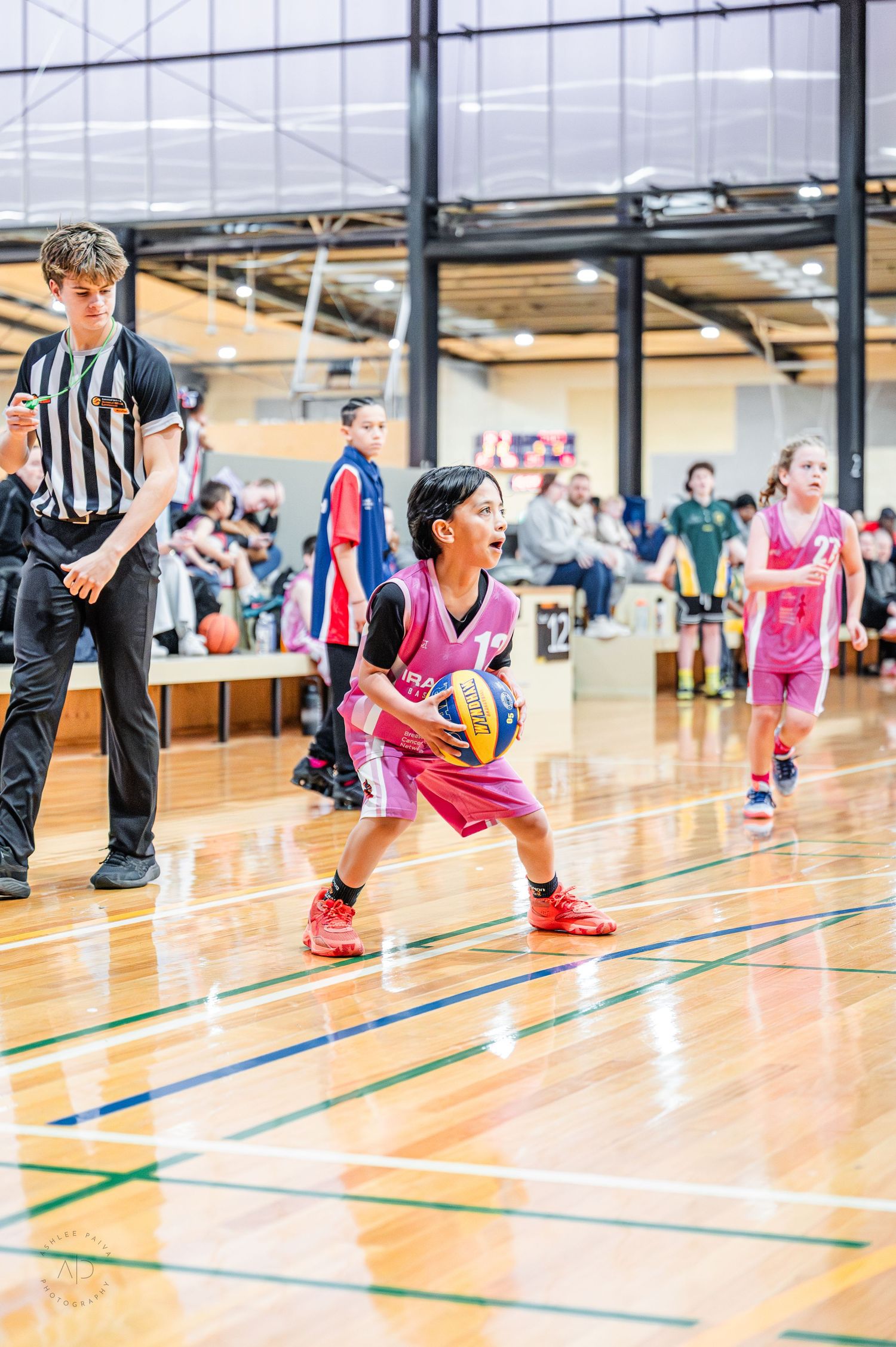 Young girl in pink uniform holds basketball during indoor youth basketball game with referee and players in background.