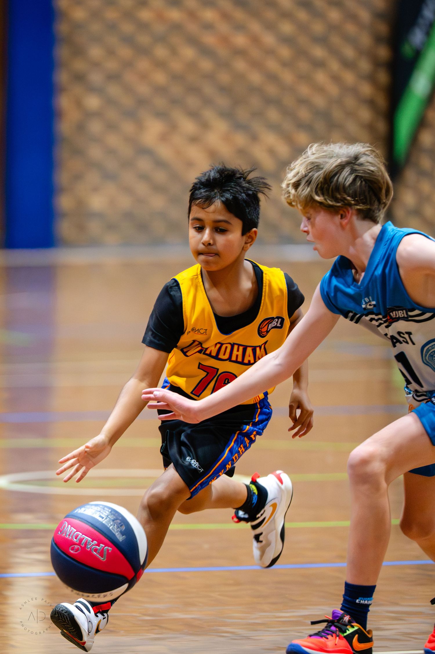 Two young boys playing basketball in a youth league game, one dribbling in a yellow jersey, the other defending in blue.