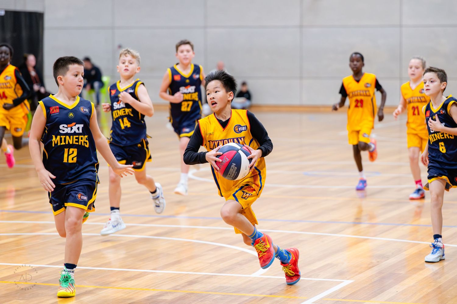Young boys playing competitive indoor basketball game, wearing yellow and navy SIXT Miners uniforms on hardwood court.
