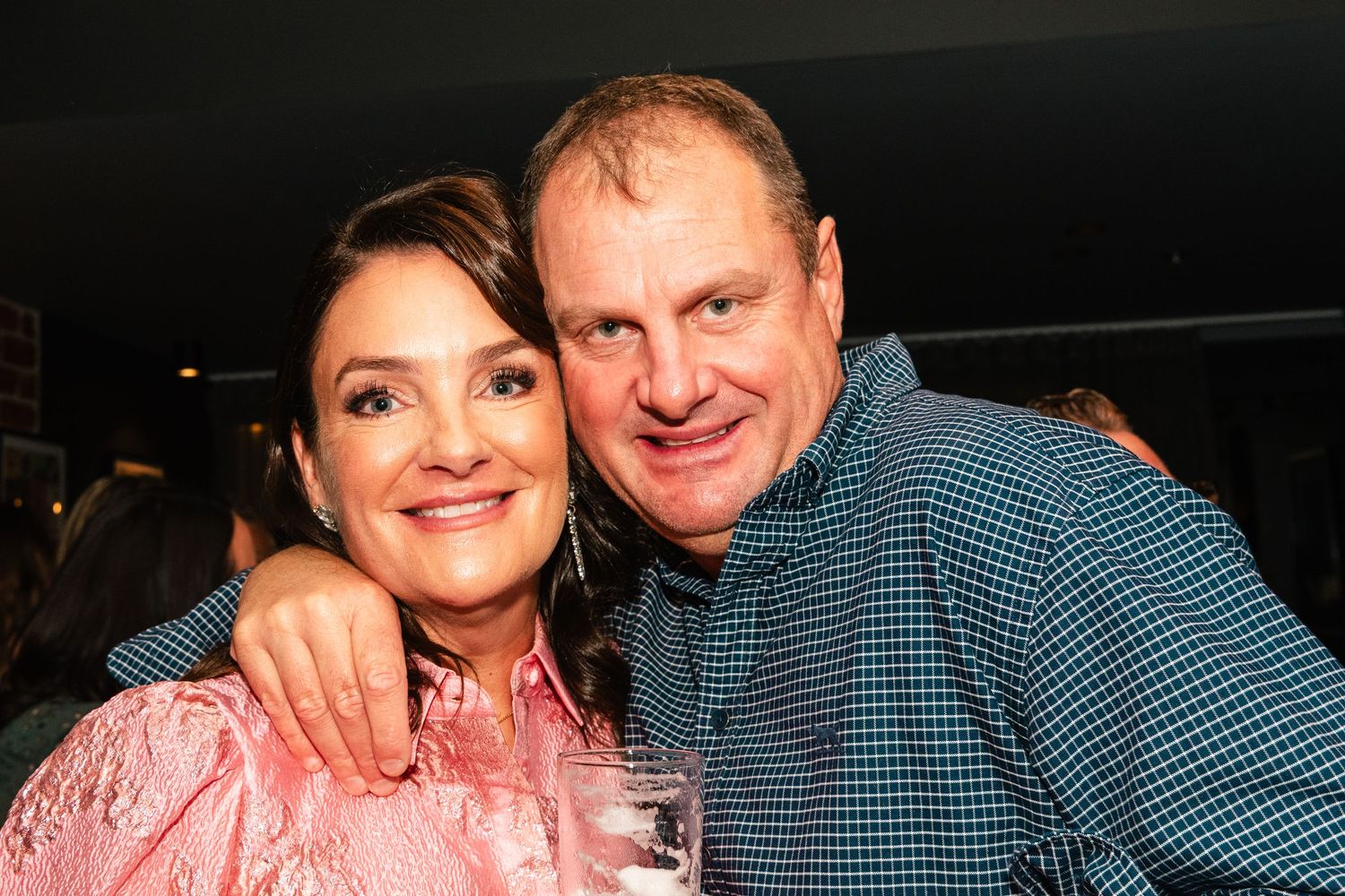 A smiling man and woman posing together at a social event, the woman in pink and the man in a blue checkered shirt.