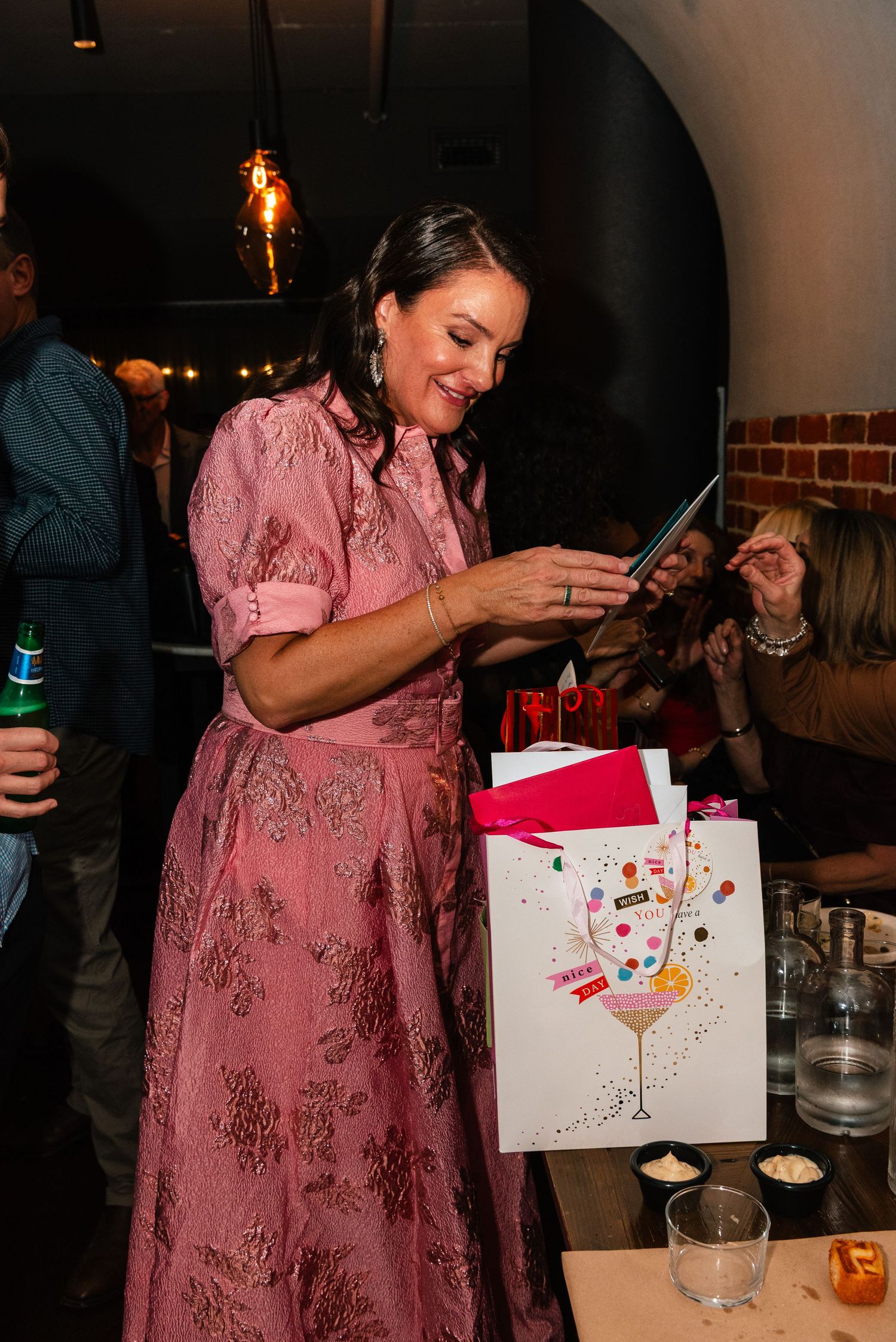 Woman in pink floral dress smiling at birthday gift bag with cocktail design at indoor party celebration.