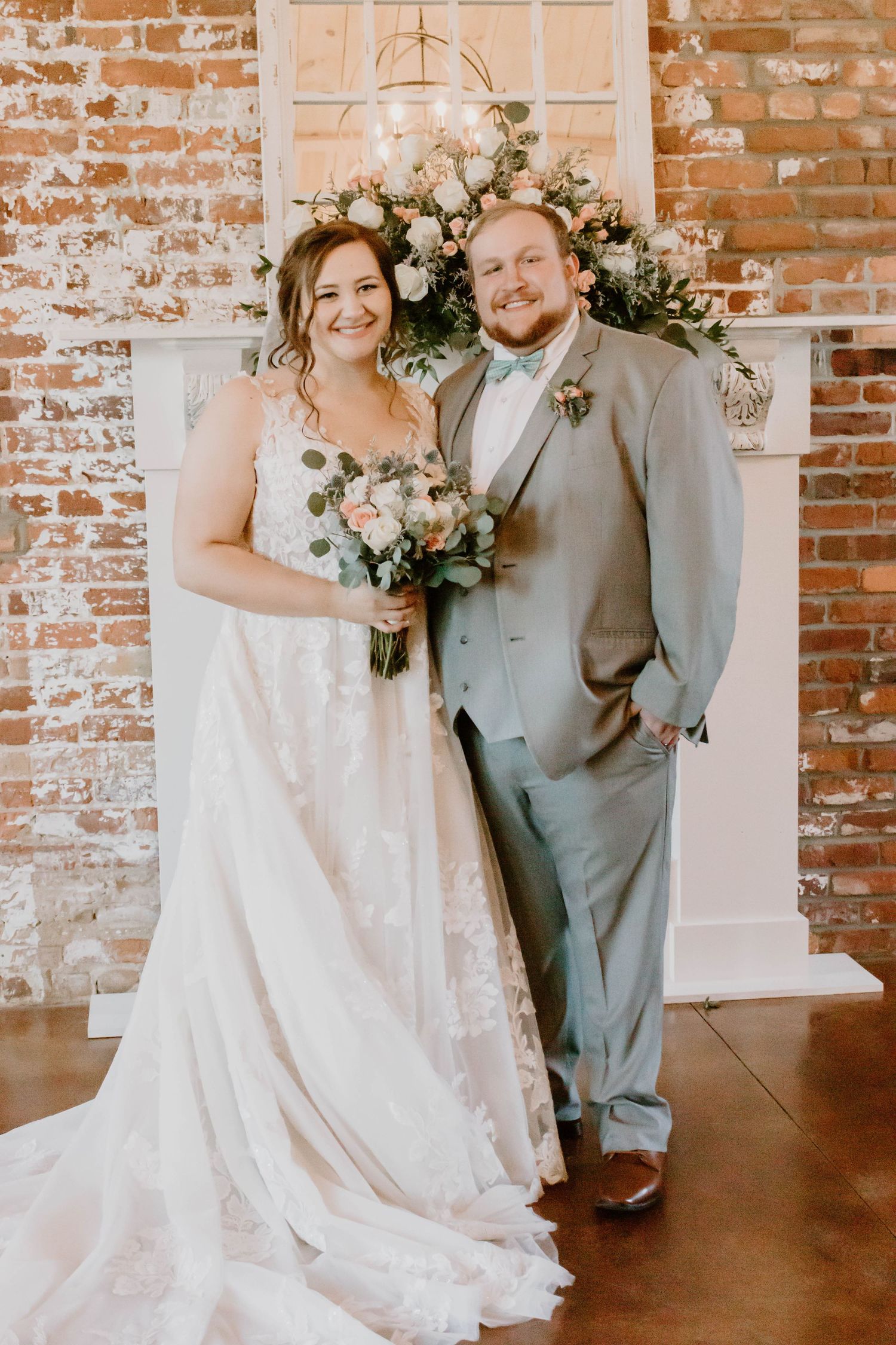 Wedding couple poses against exposed brick wall with floral arrangements and elegant decor.