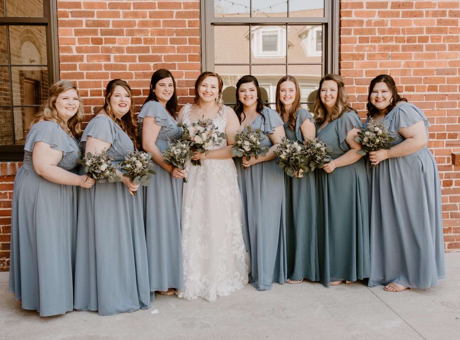 Wedding party in dusty blue bridesmaid dresses and white bridal gown pose against brick wall.