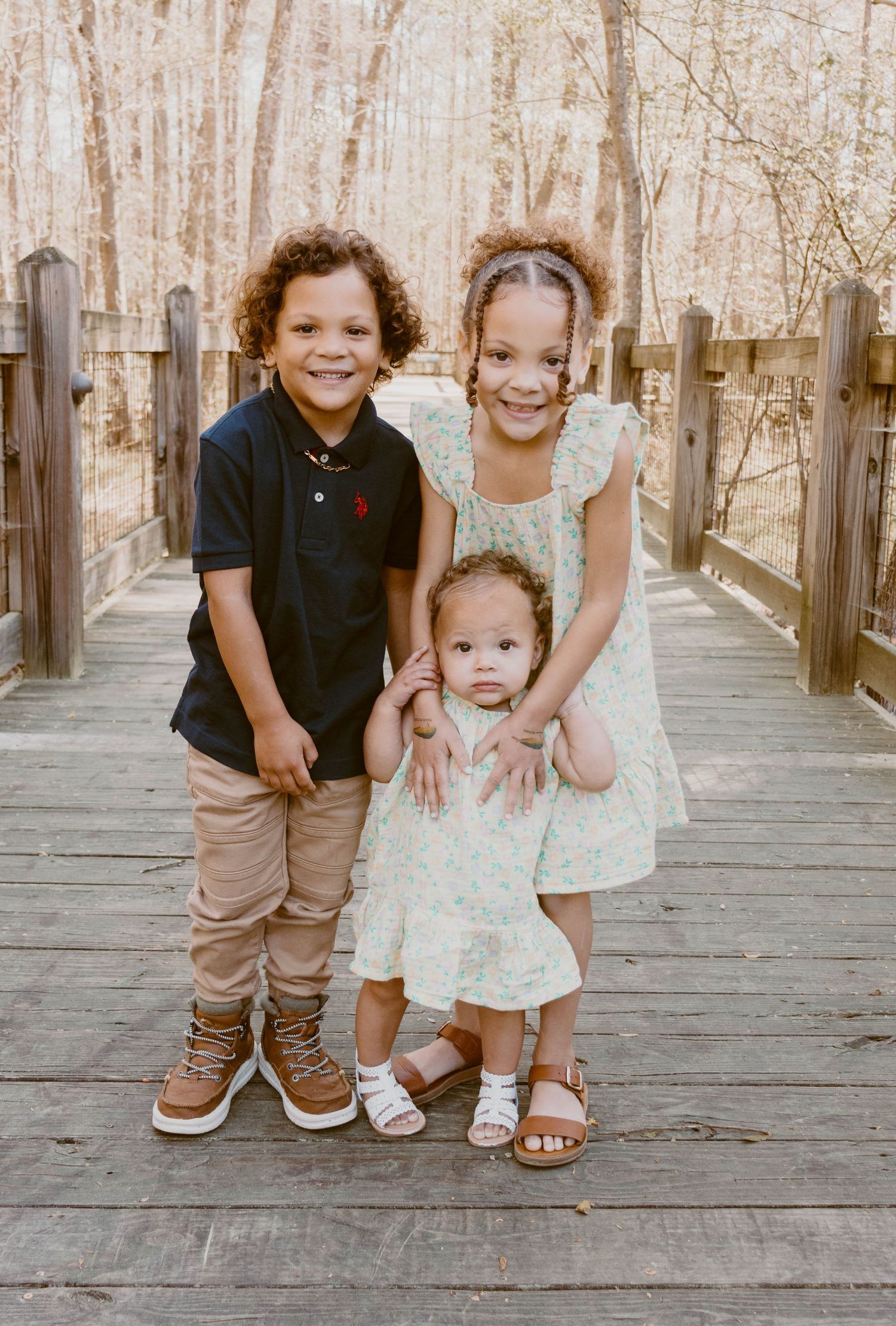 Three siblings pose together on a wooden boardwalk surrounded by trees in a natural setting.