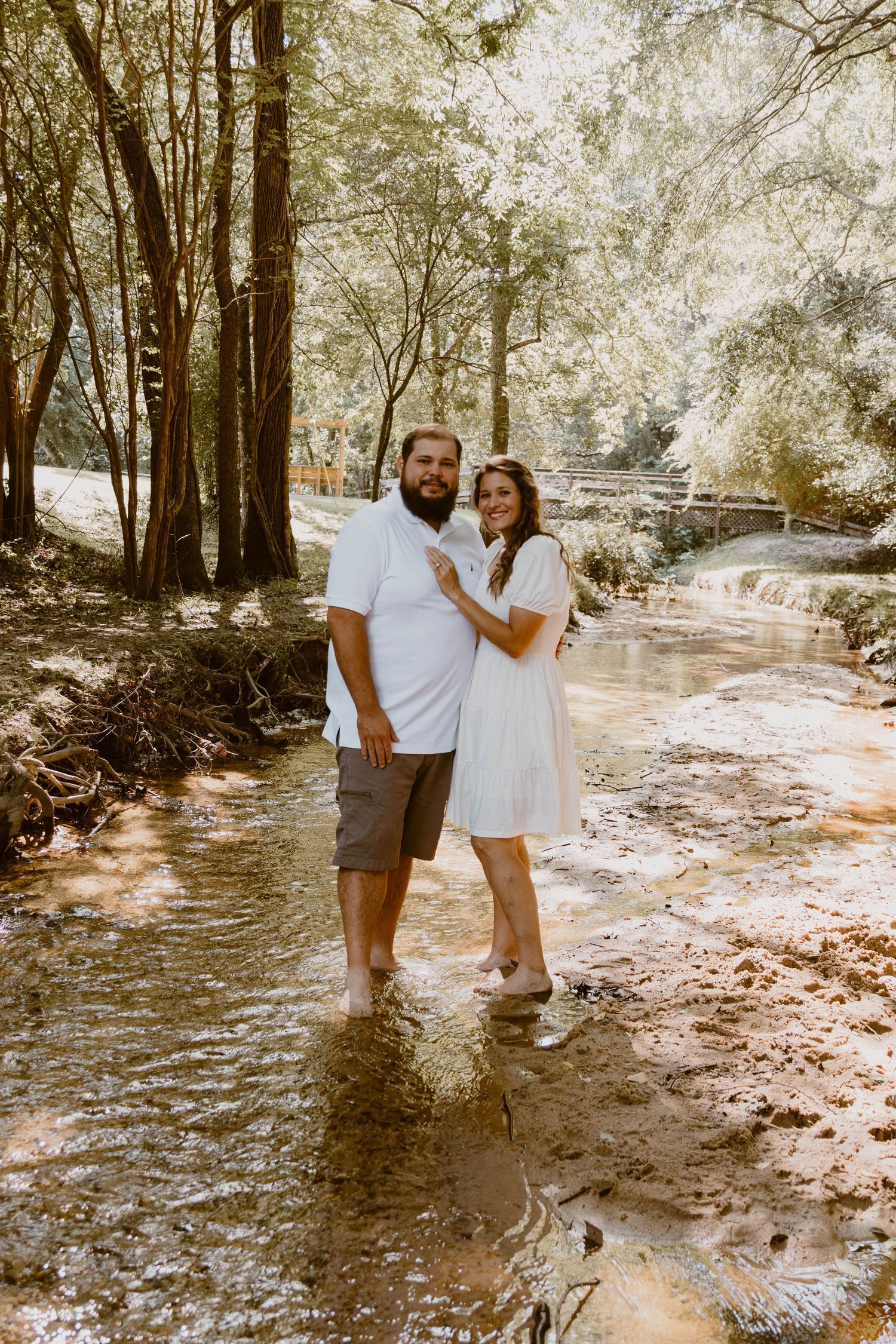 A couple stands together in a creek bed surrounded by trees during a sunny outdoor photo session.