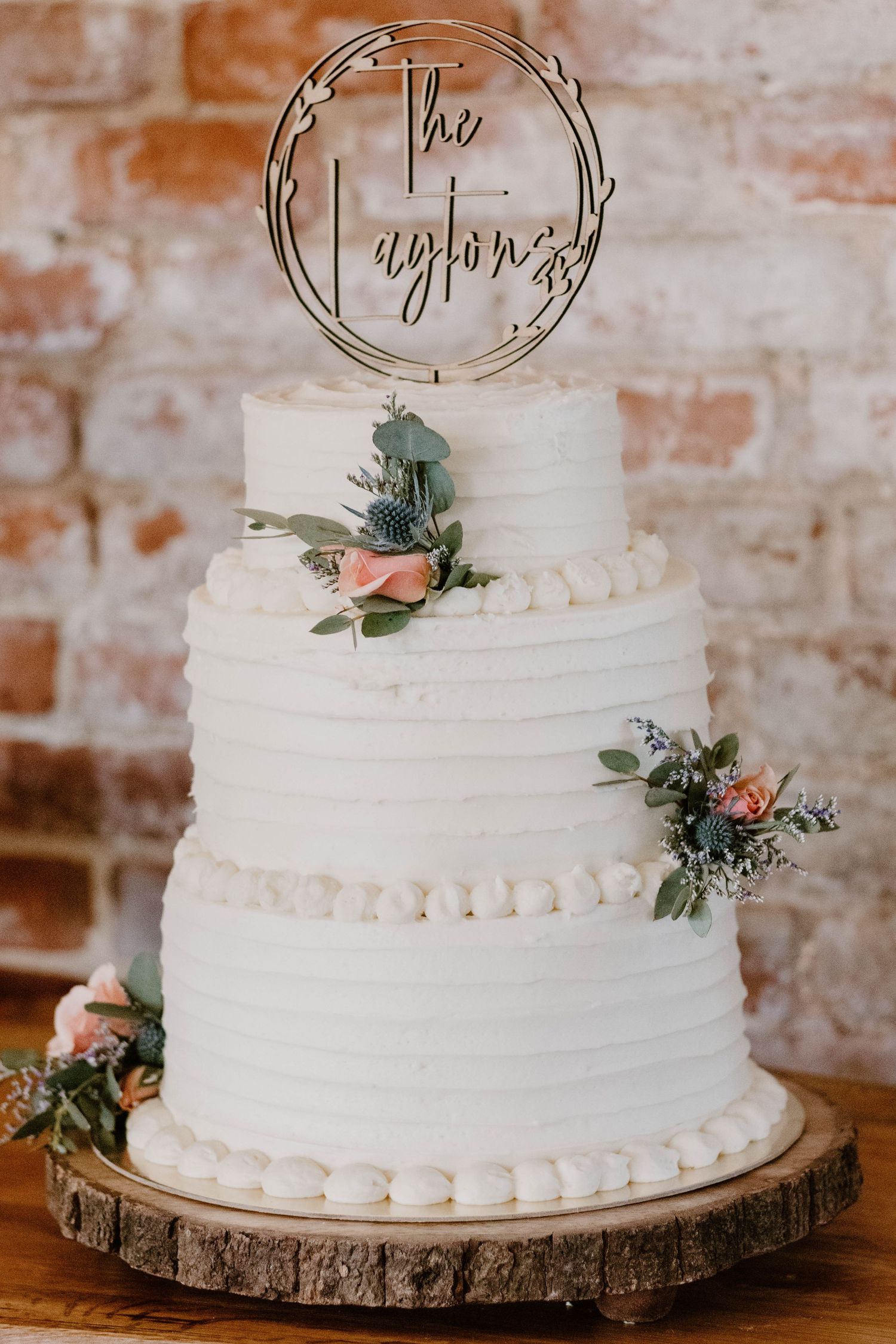 White tiered wedding cake with metallic topper displayed on rustic wood slice against brick wall.
