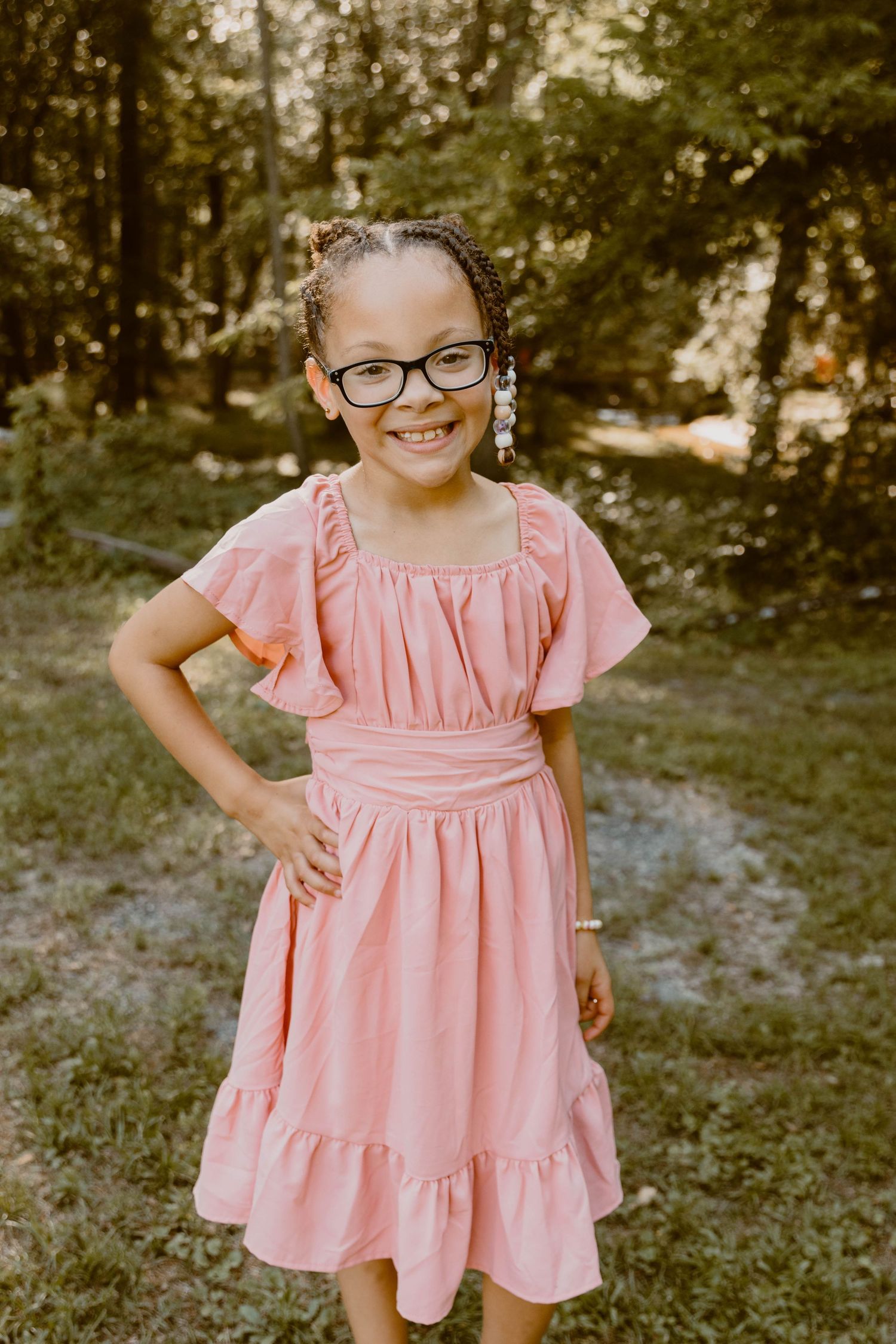 A young child in a pink ruffle dress and glasses poses confidently outdoors.
