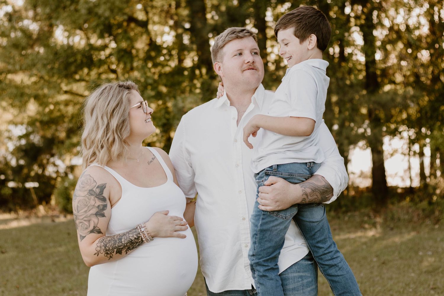 A family maternity photoshoot in a wooded setting features a couple and their young child.