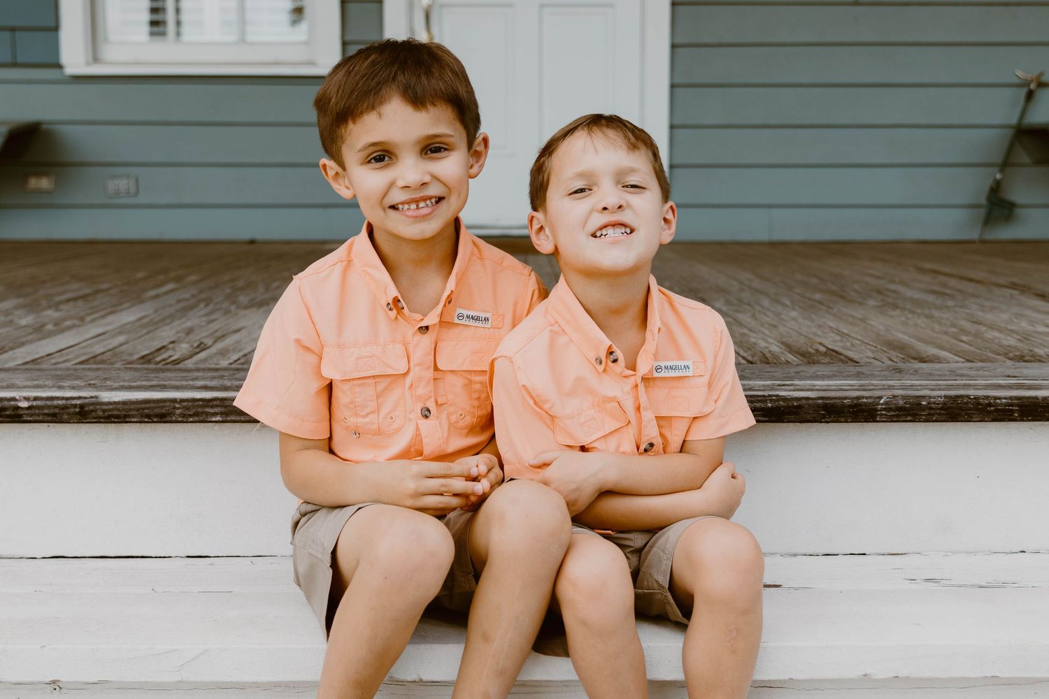 Two children in matching coral shirts sitting on white porch steps.