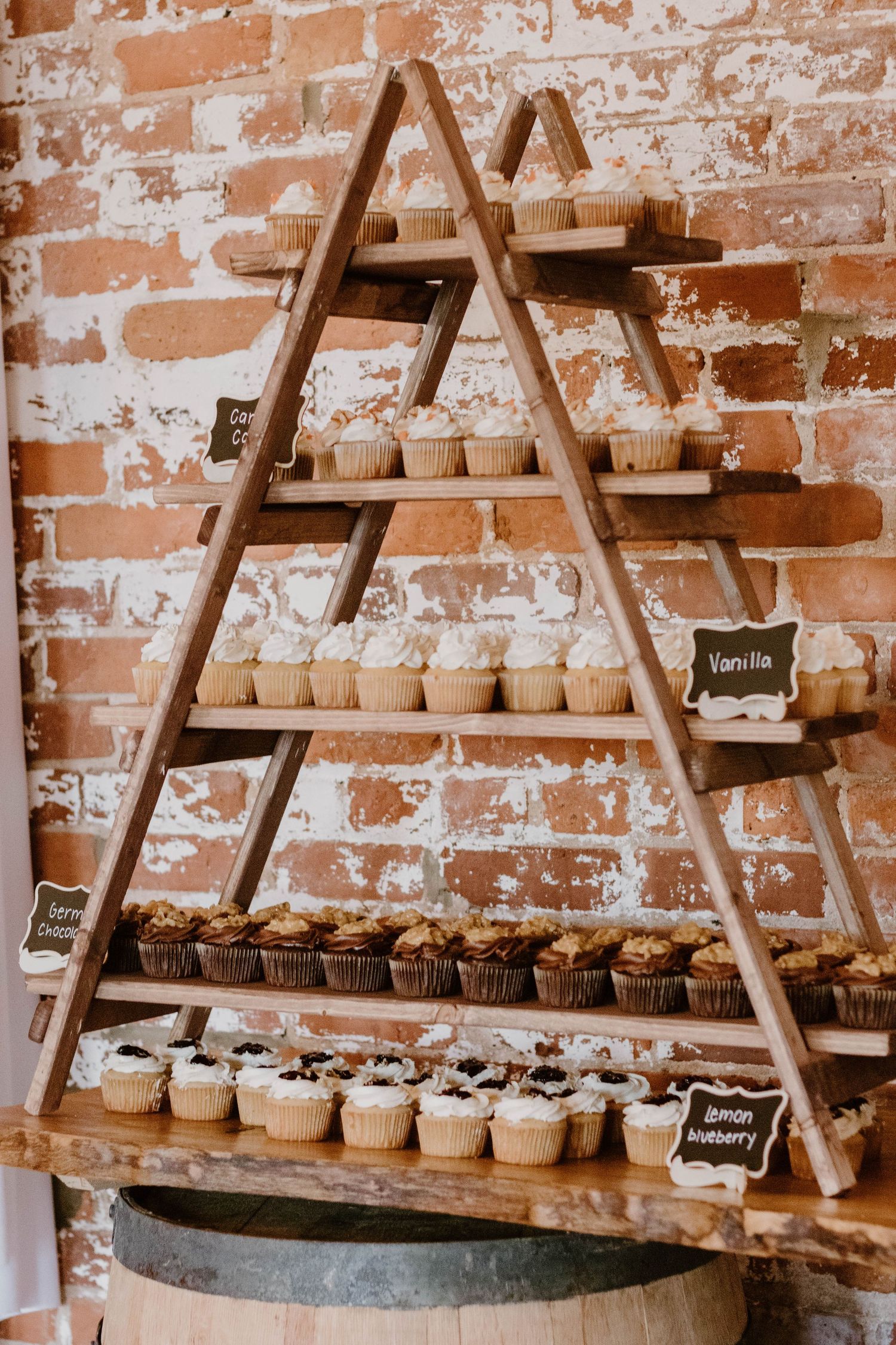 Rustic wooden ladder display with tiers of cupcakes against exposed brick wall.