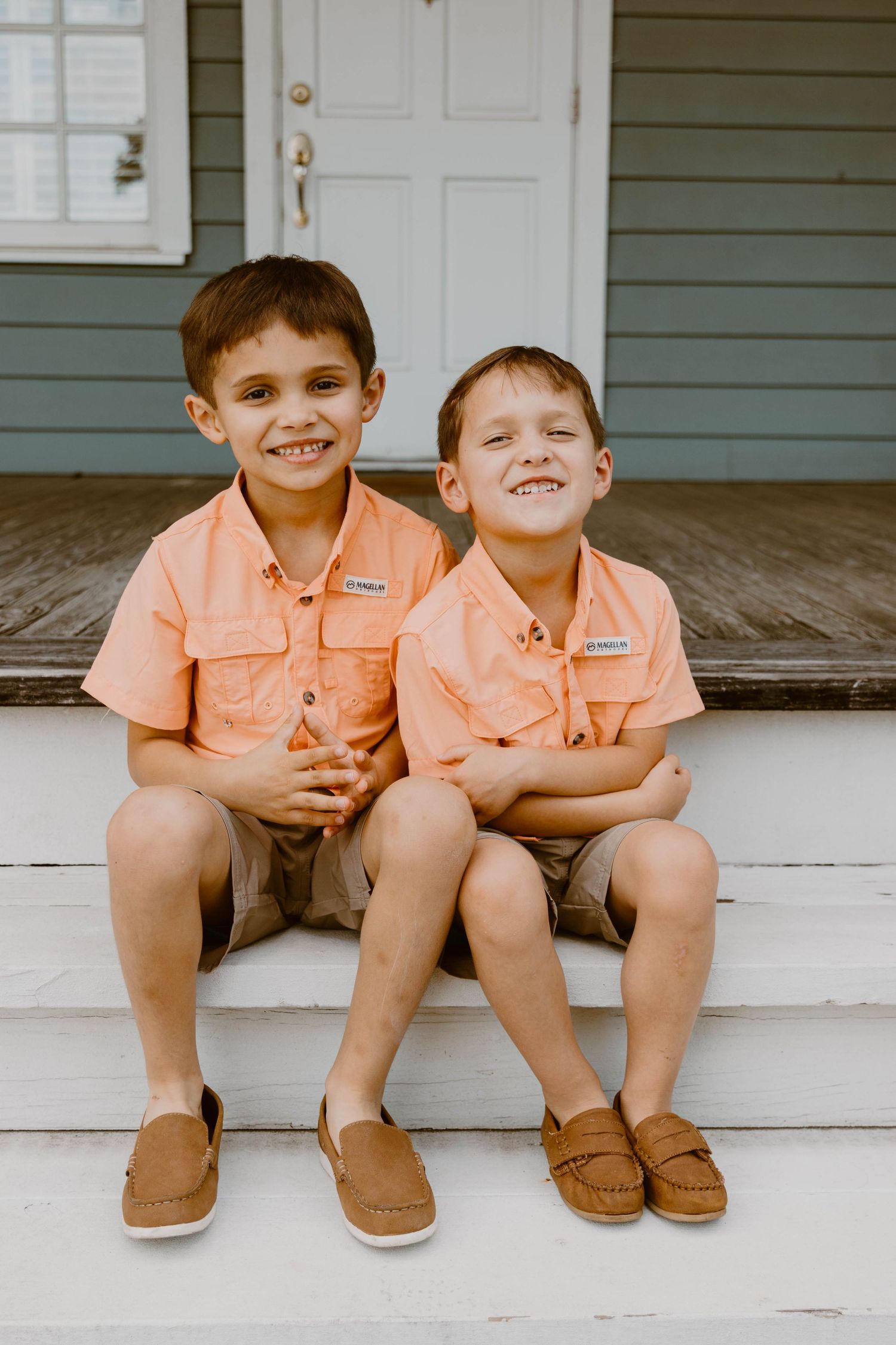 Two young boys in matching coral shirts smiling on white porch steps.