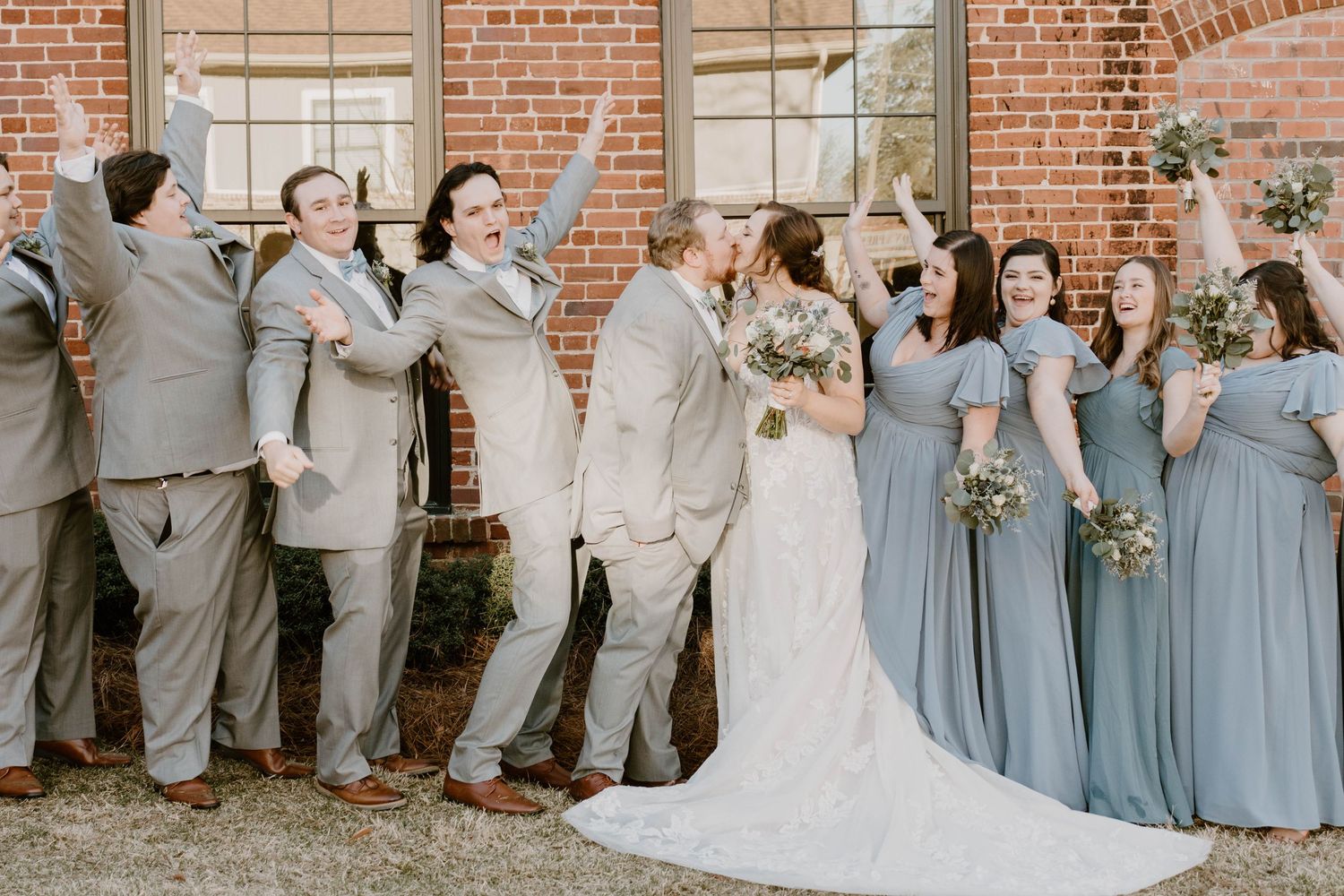 Wedding party in grey and dusty blue attire poses against brick building exterior.