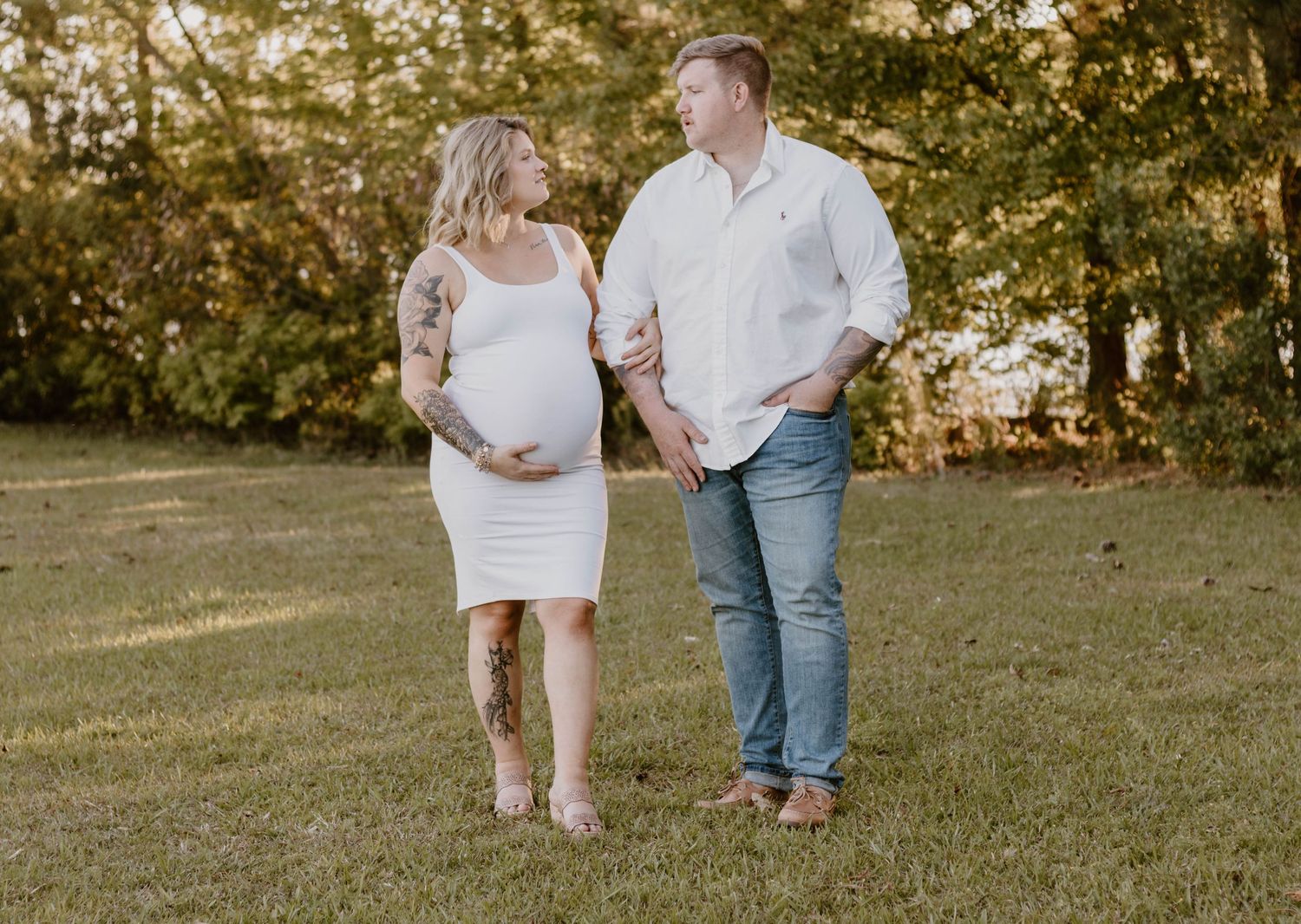A maternity photo session in a sunlit field shows an expectant couple walking together in casual attire.