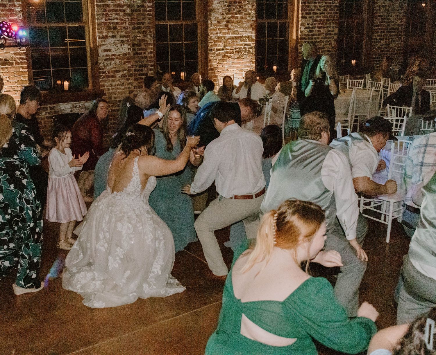 Wedding guests dance and celebrate on the dance floor at a rustic brick venue with white chairs.