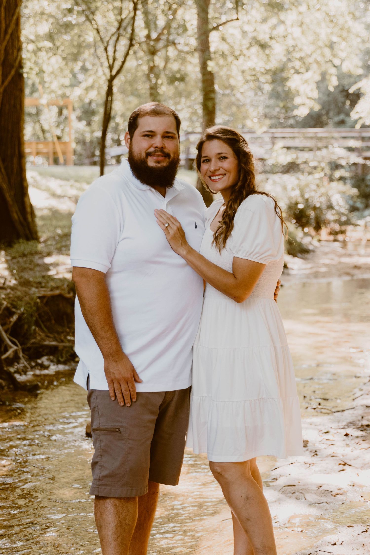 A couple poses together in a romantic outdoor photoshoot by a stream in a sunlit wooded area.