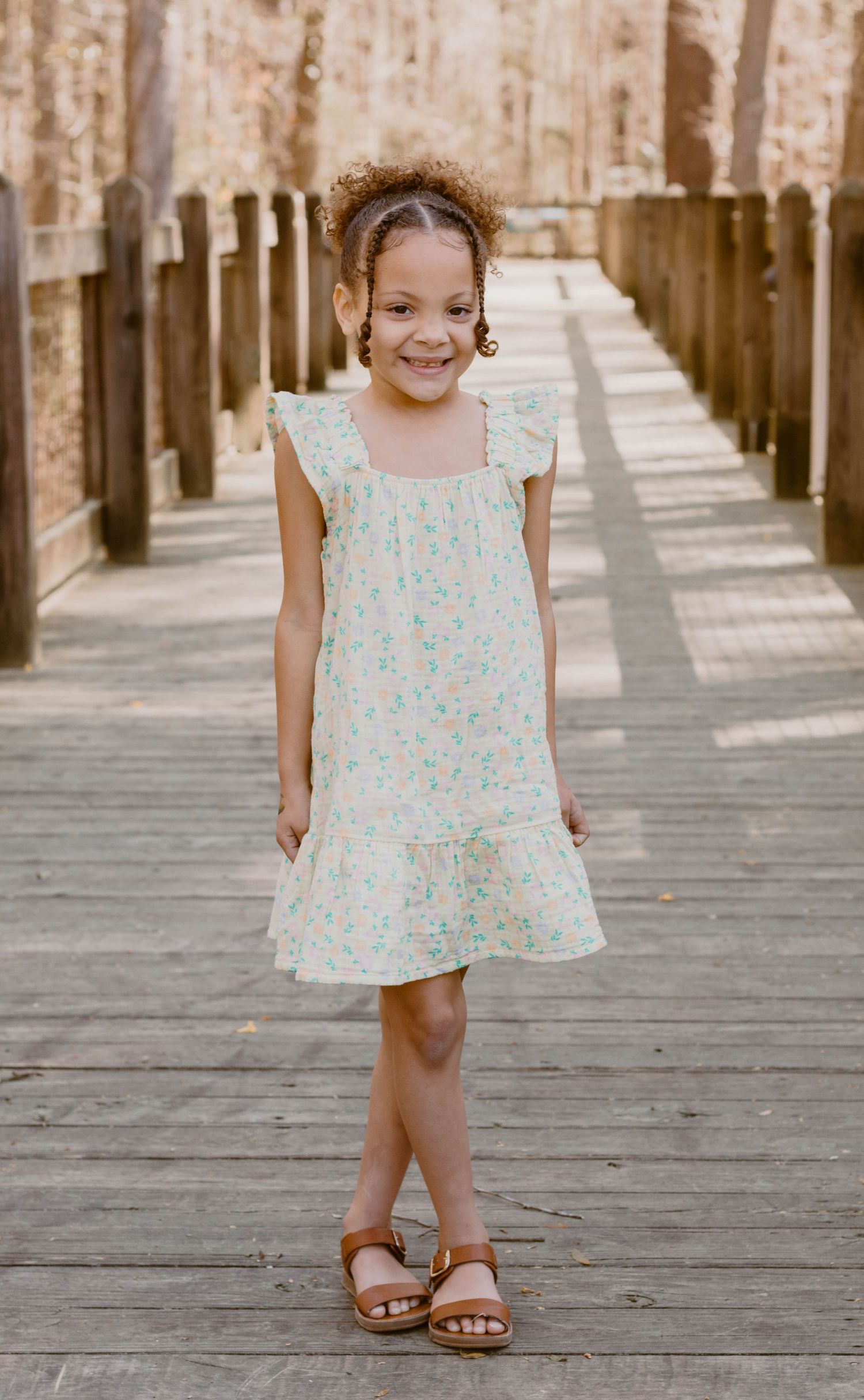 A child in a floral dress poses on a wooden boardwalk with a bright smile.