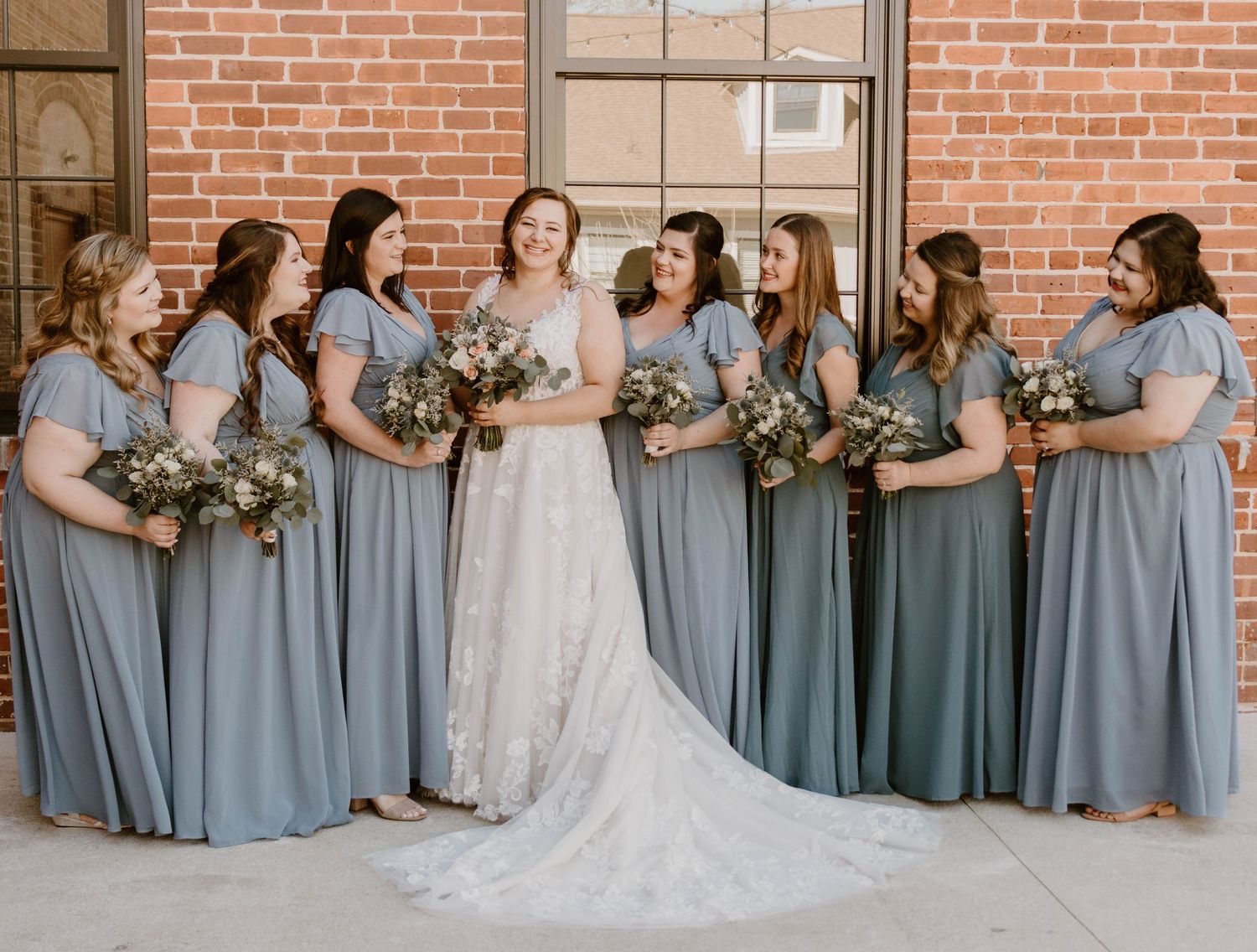 Bridal party wearing matching blue dresses lined up for formal photo against brick backdrop.