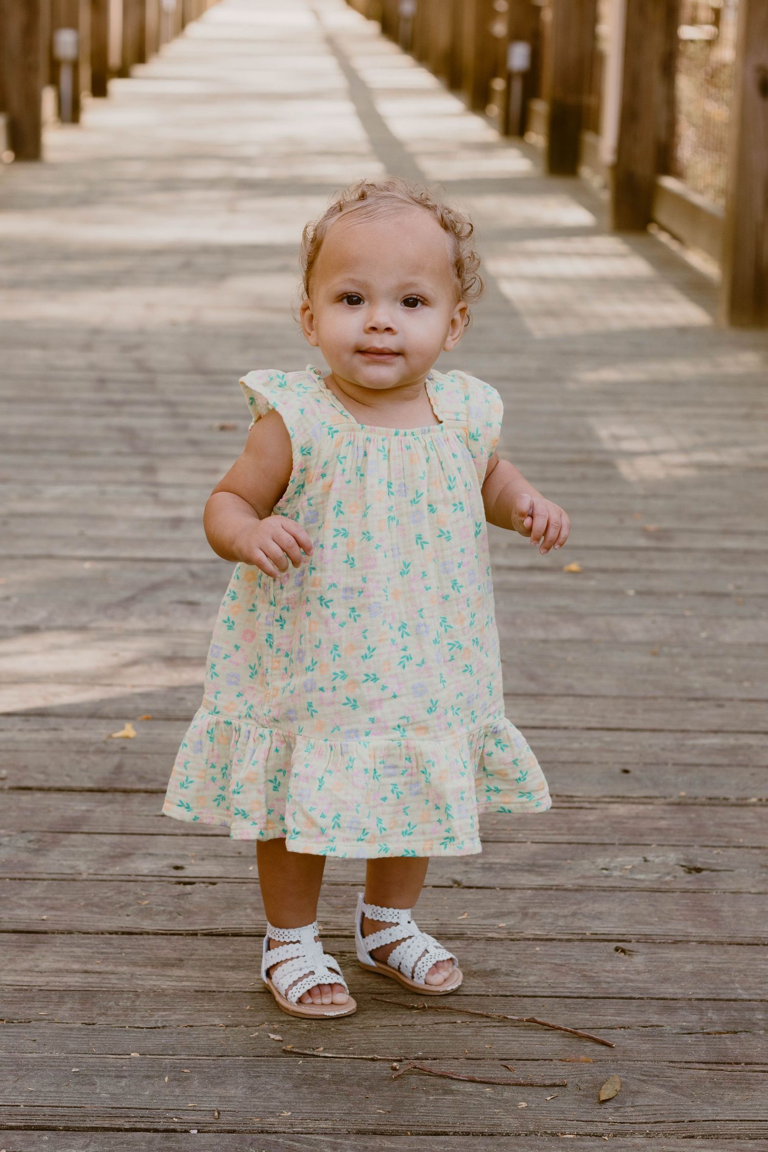 Toddler in floral dress walking on wooden boardwalk during family photo session.