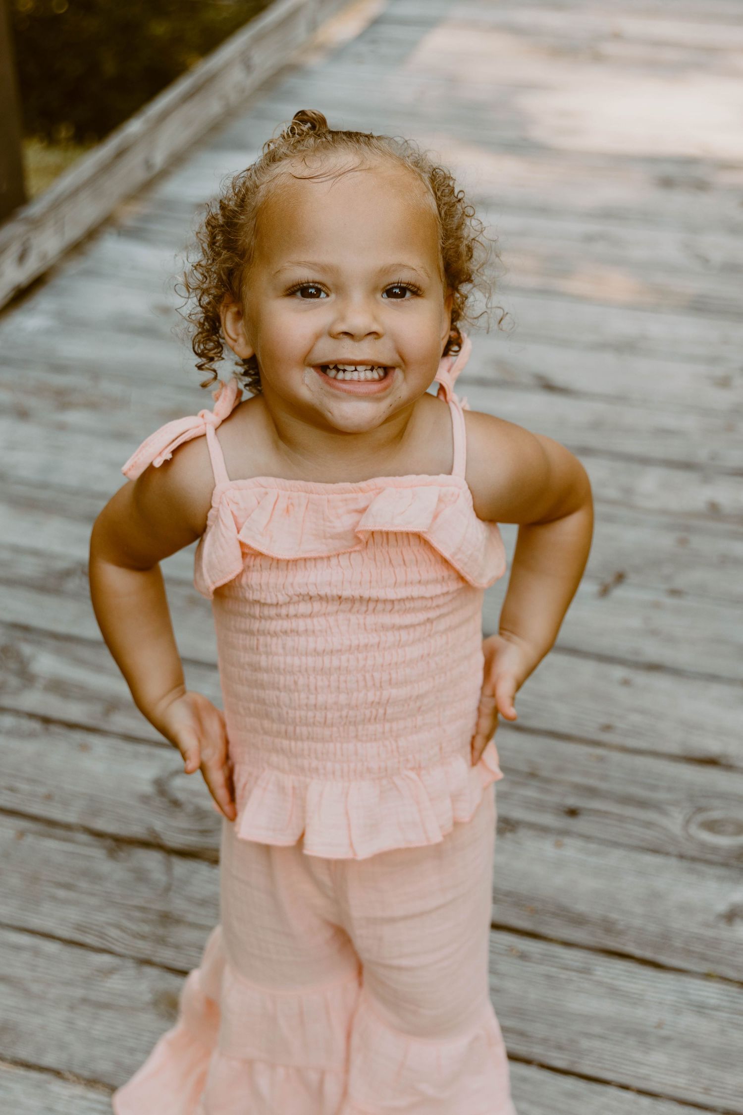 A toddler in a pink ruffled outfit smiles brightly on a wooden deck.