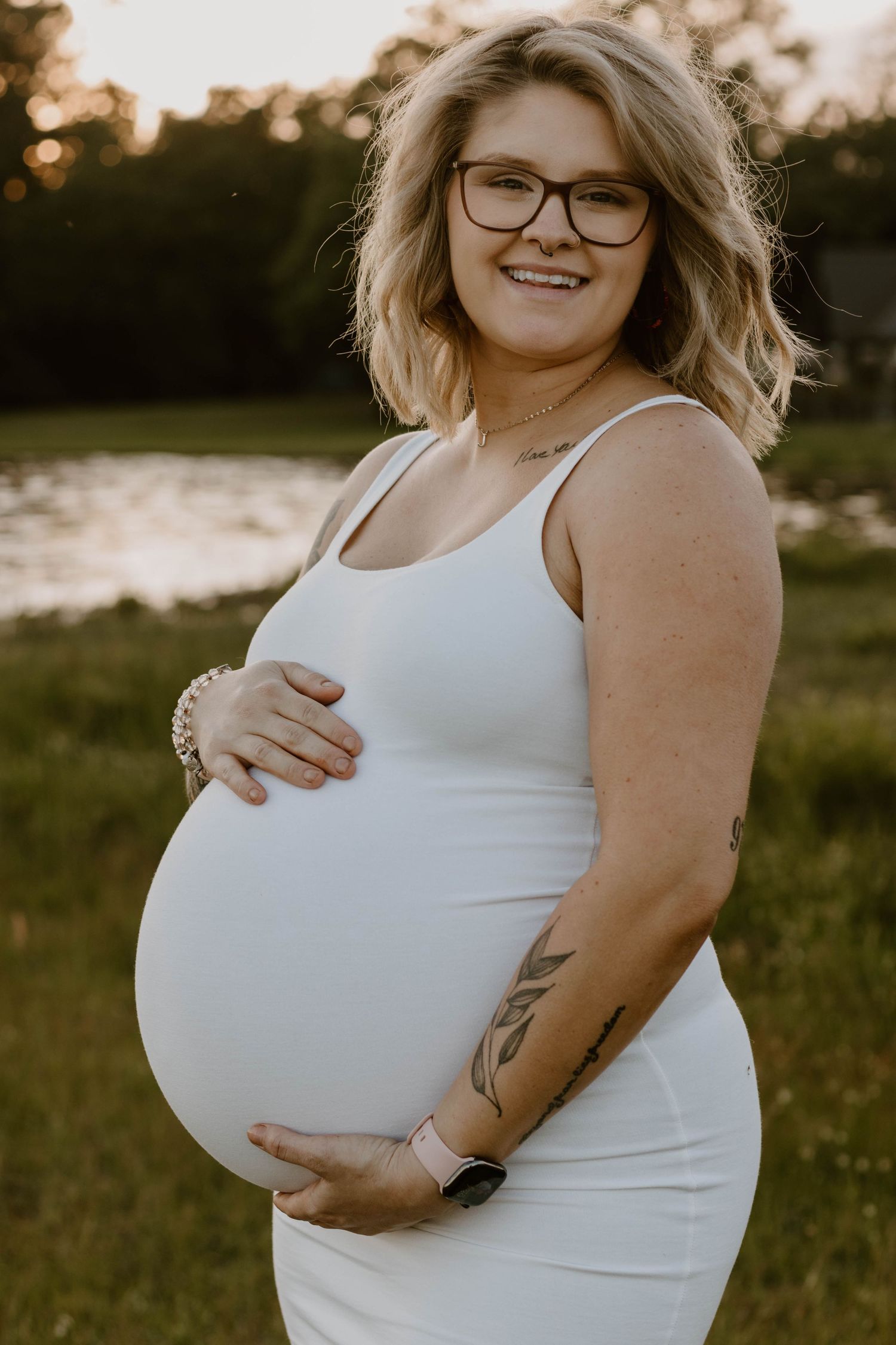 Expectant mother in white tank top poses outdoors during sunset photoshoot.