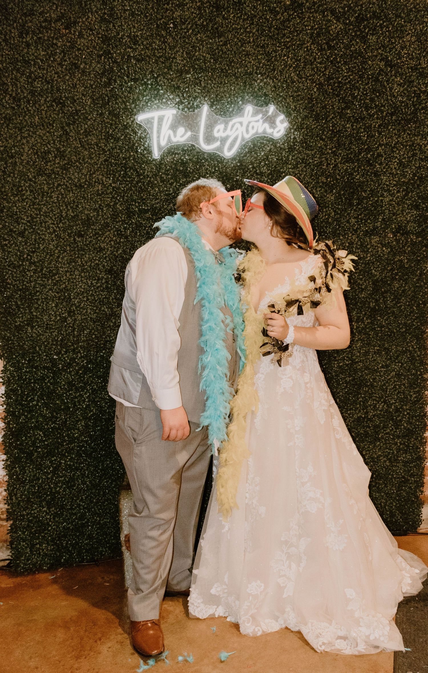 Couple shares romantic moment under neon sign wearing feather boas at wedding reception.