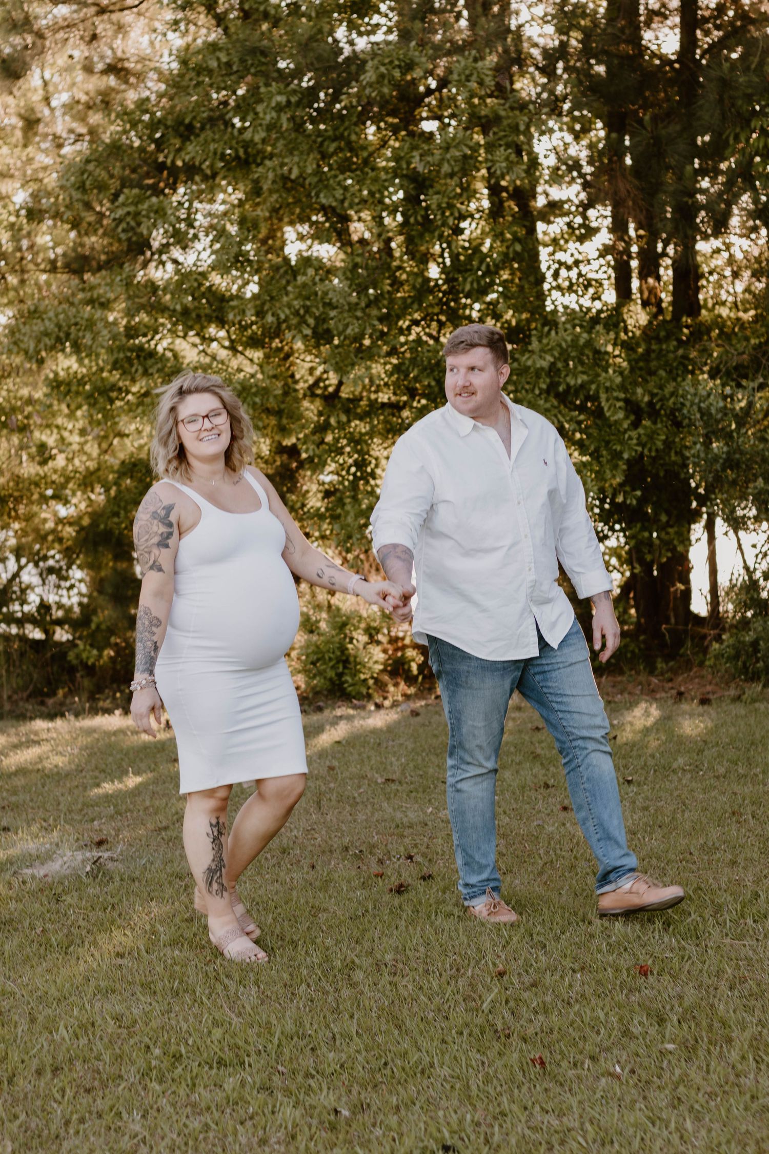An expecting couple takes a stroll through a golden-lit forest clearing during their maternity photoshoot.