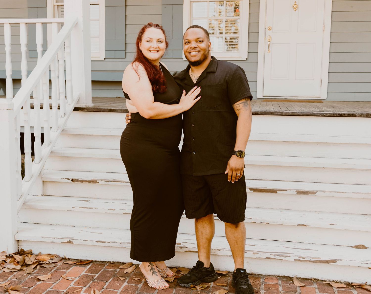A couple poses together on white porch steps wearing matching black outfits.