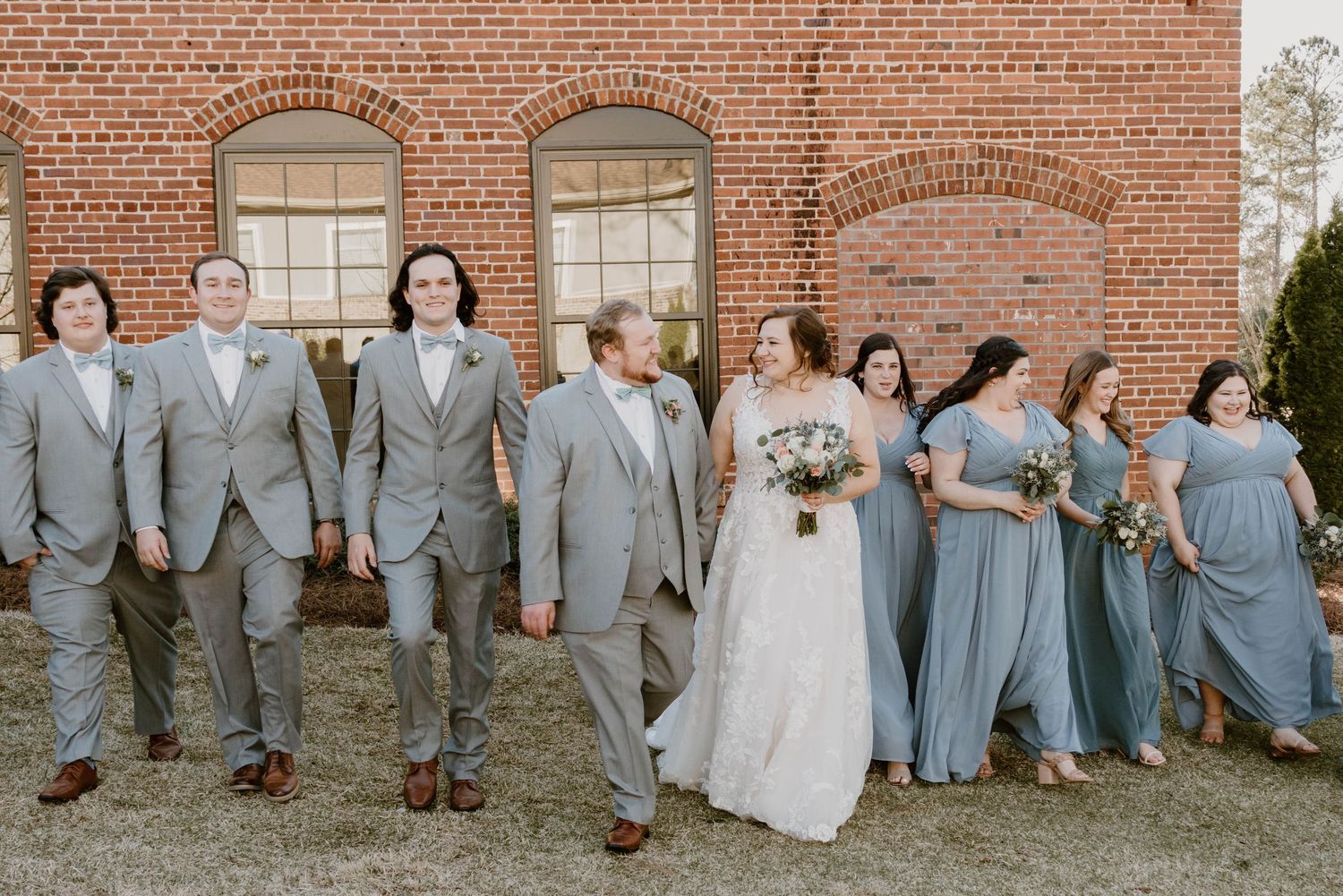 Wedding party members arranged along brick wall in coordinated grey and blue attire.