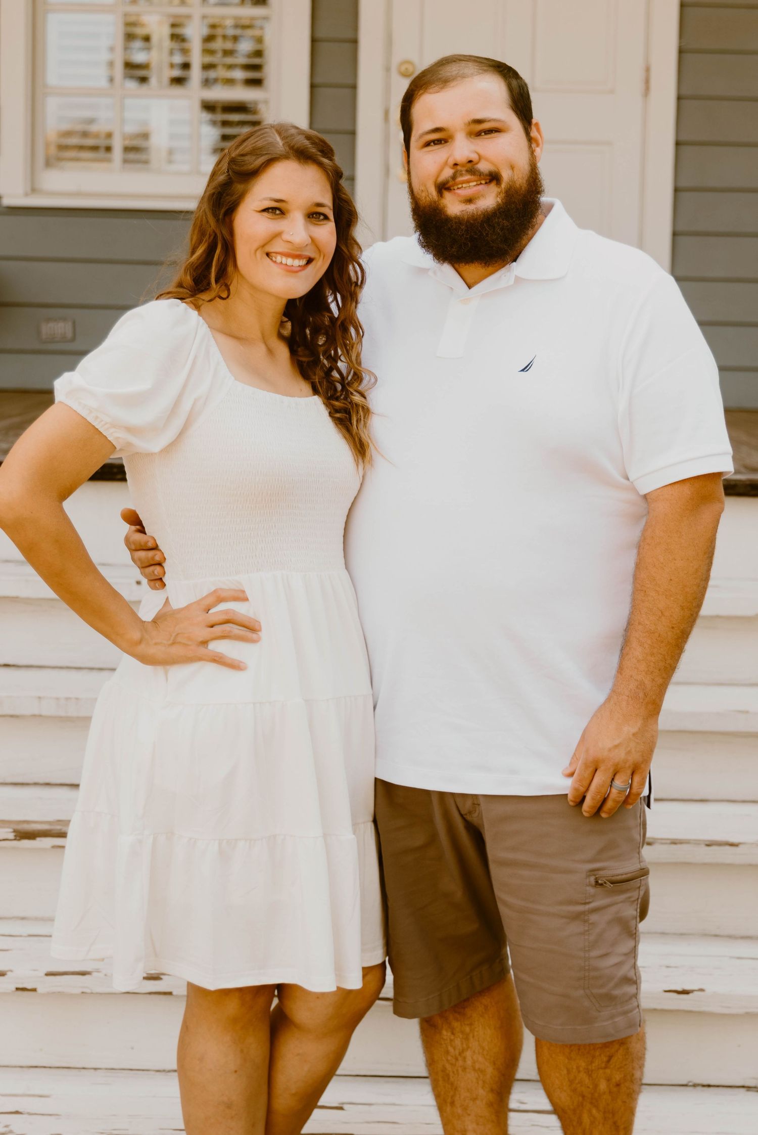 Couple in white shirts posing casually on porch of gray house.