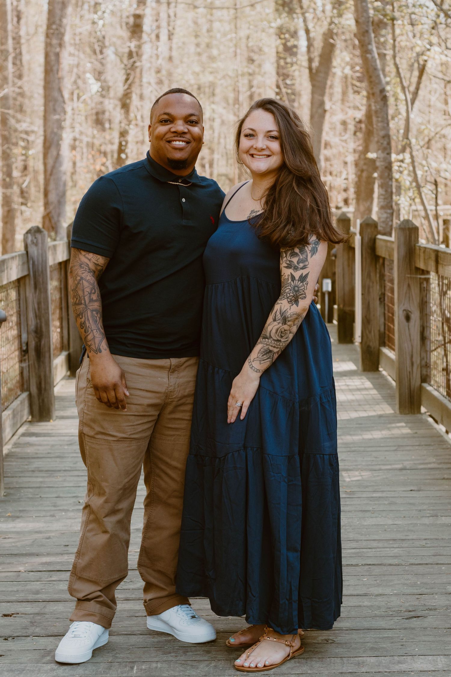 Couple poses together on a rustic wooden boardwalk surrounded by trees in casual attire.