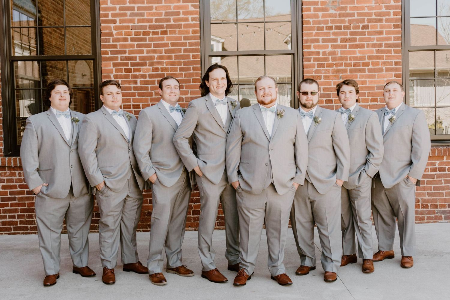 Group photo of wedding party members in matching light gray suits against brick building.