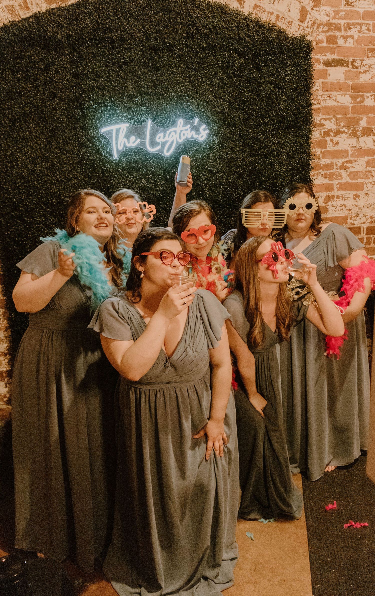 Bridesmaids in matching gray dresses pose with colorful boas in front of a neon sign.