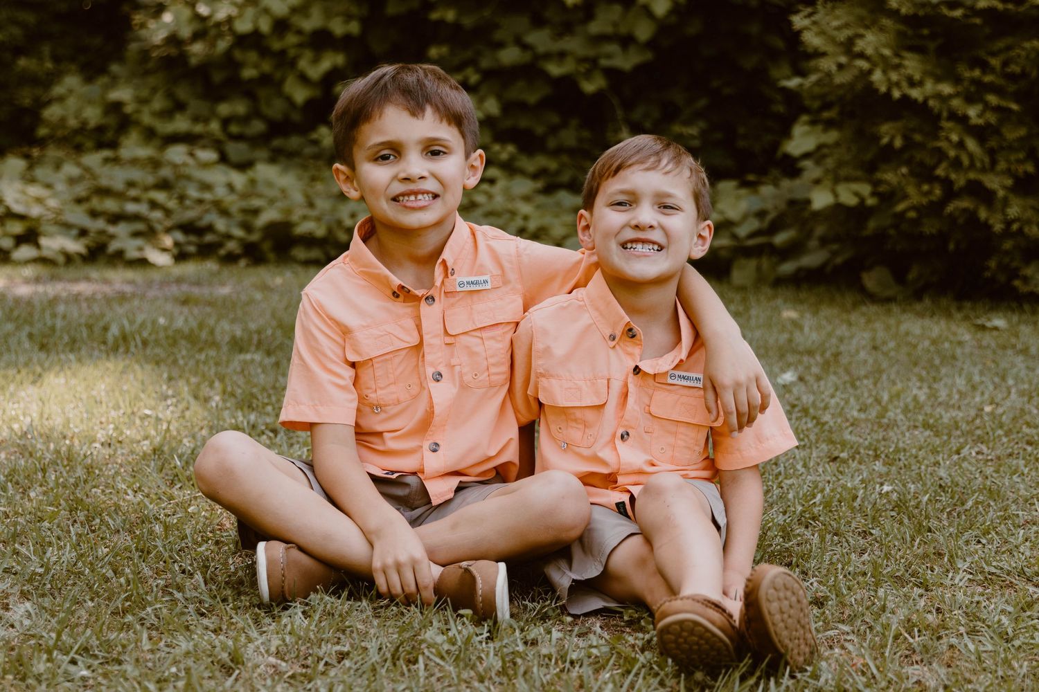 Children sitting together on grass wearing matching peach-colored outfits.