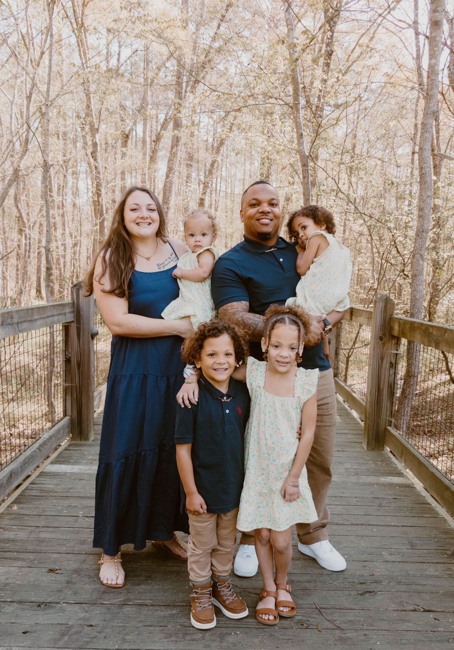 A family of six stands together on a wooden walkway in a wooded setting.