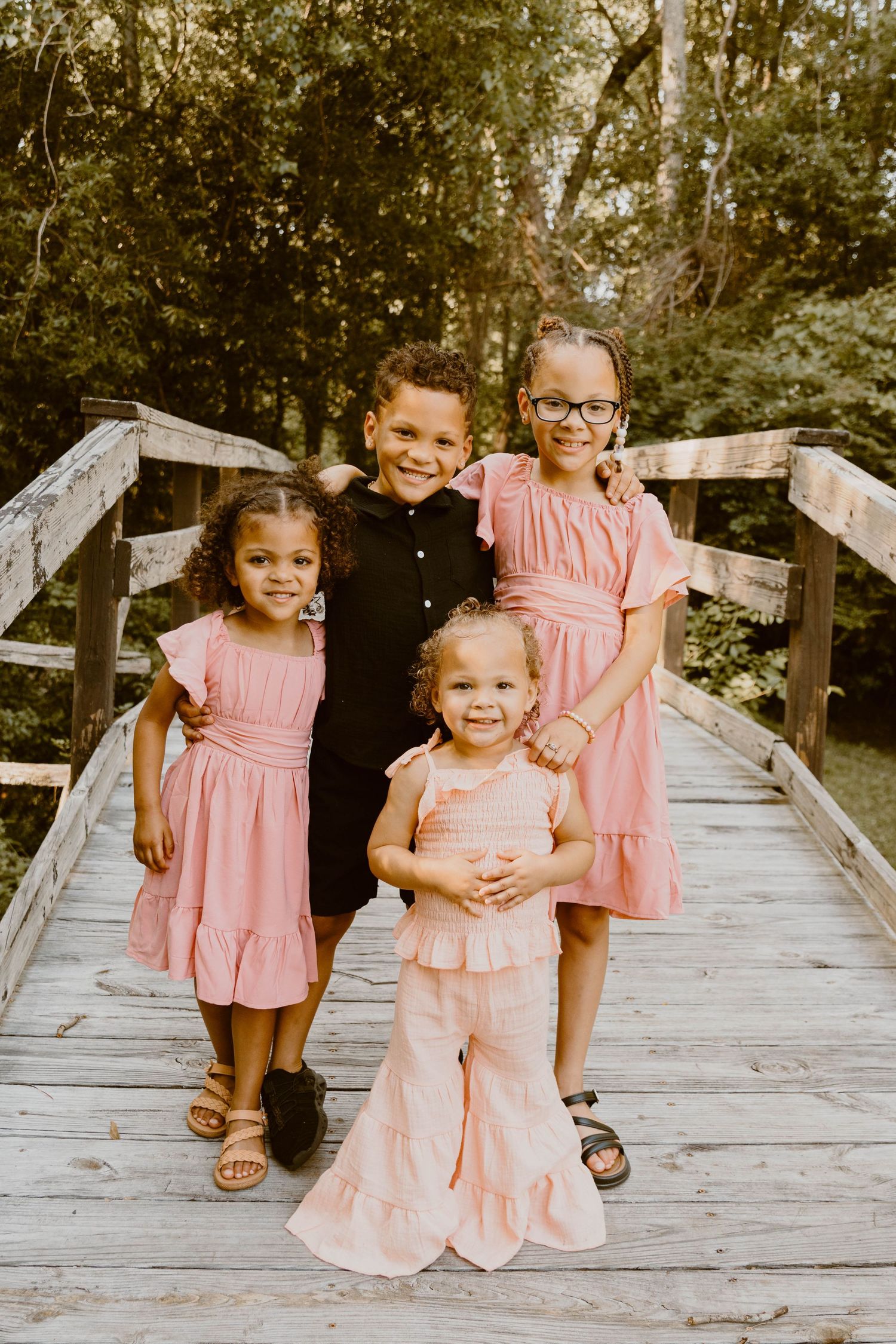 Four children in coordinating pink and black outfits pose together on a wooden bridge.